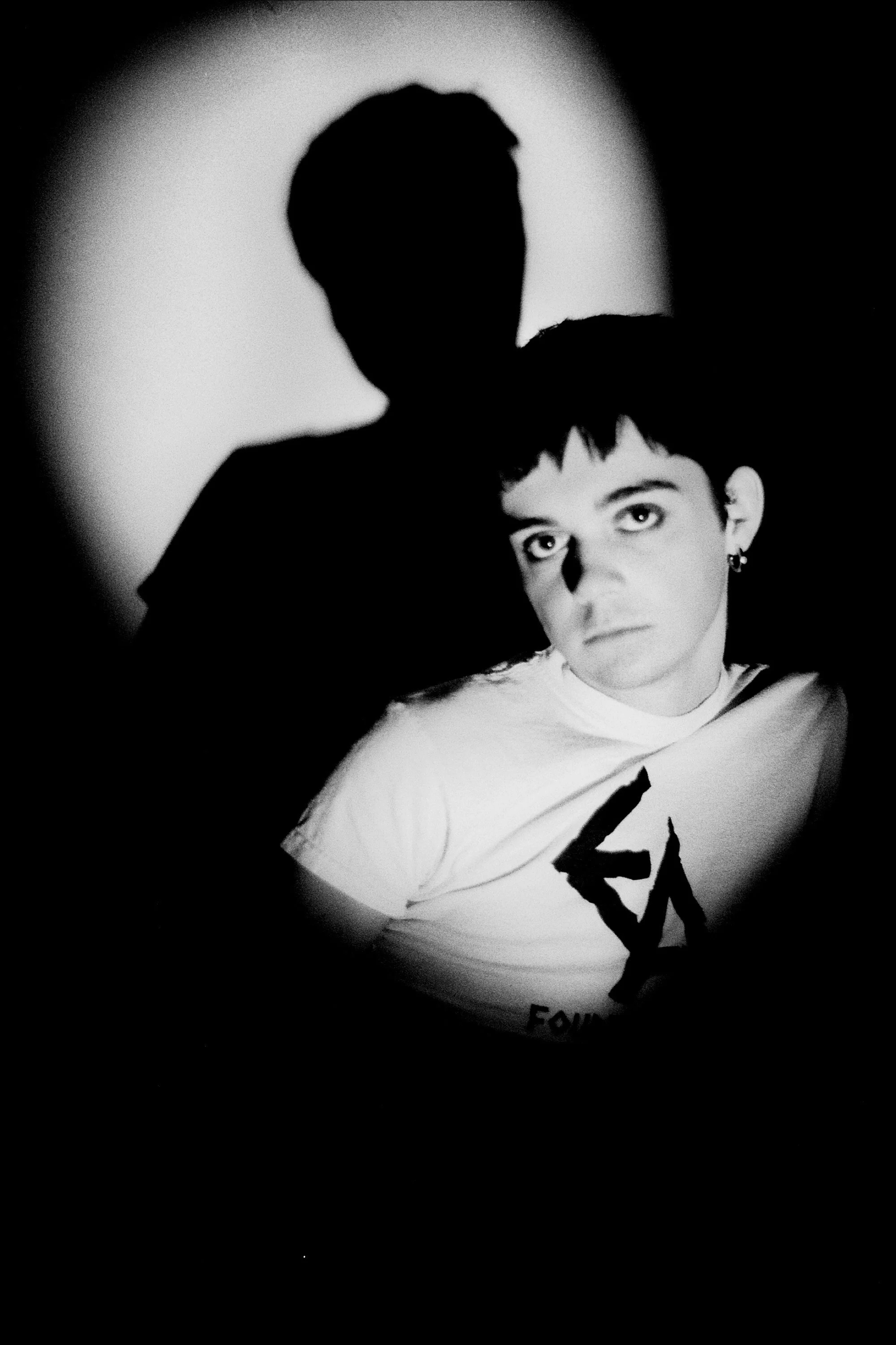 A black and white photo of a young person with short hair and earrings, looking directly at the camera, with a shadow of a person behind and above them, cast on the wall.