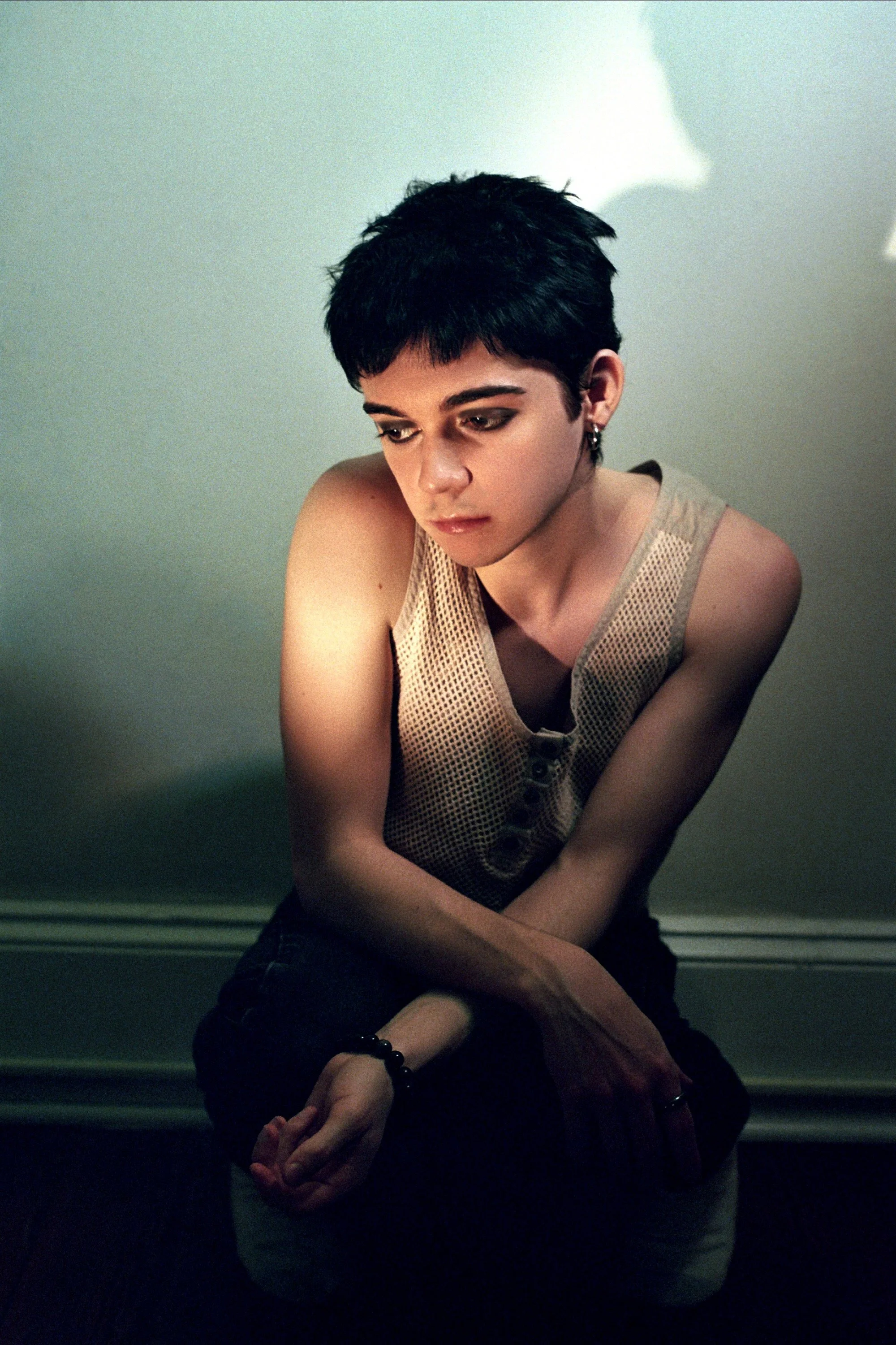 A young person with short black hair, wearing a sleeveless mesh shirt, sitting on the floor with arms crossed and looking down, against a plain wall background.