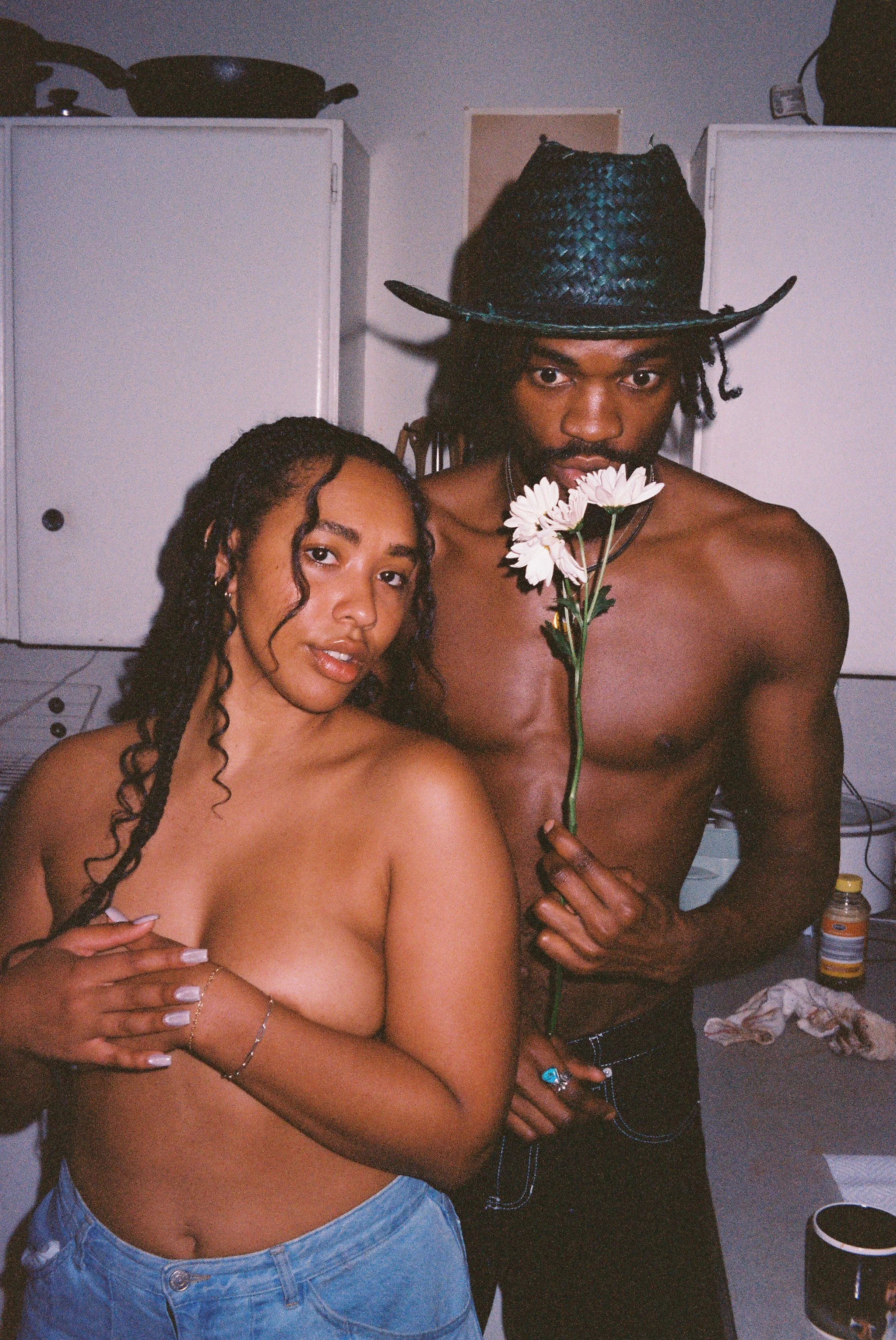 A topless woman and a topless man, with the man wearing a large black cowboy hat, stand close together. The woman has long curly hair and tattoos on her arm, and is holding her chest with one hand. The man is holding a bunch of white daisies close to