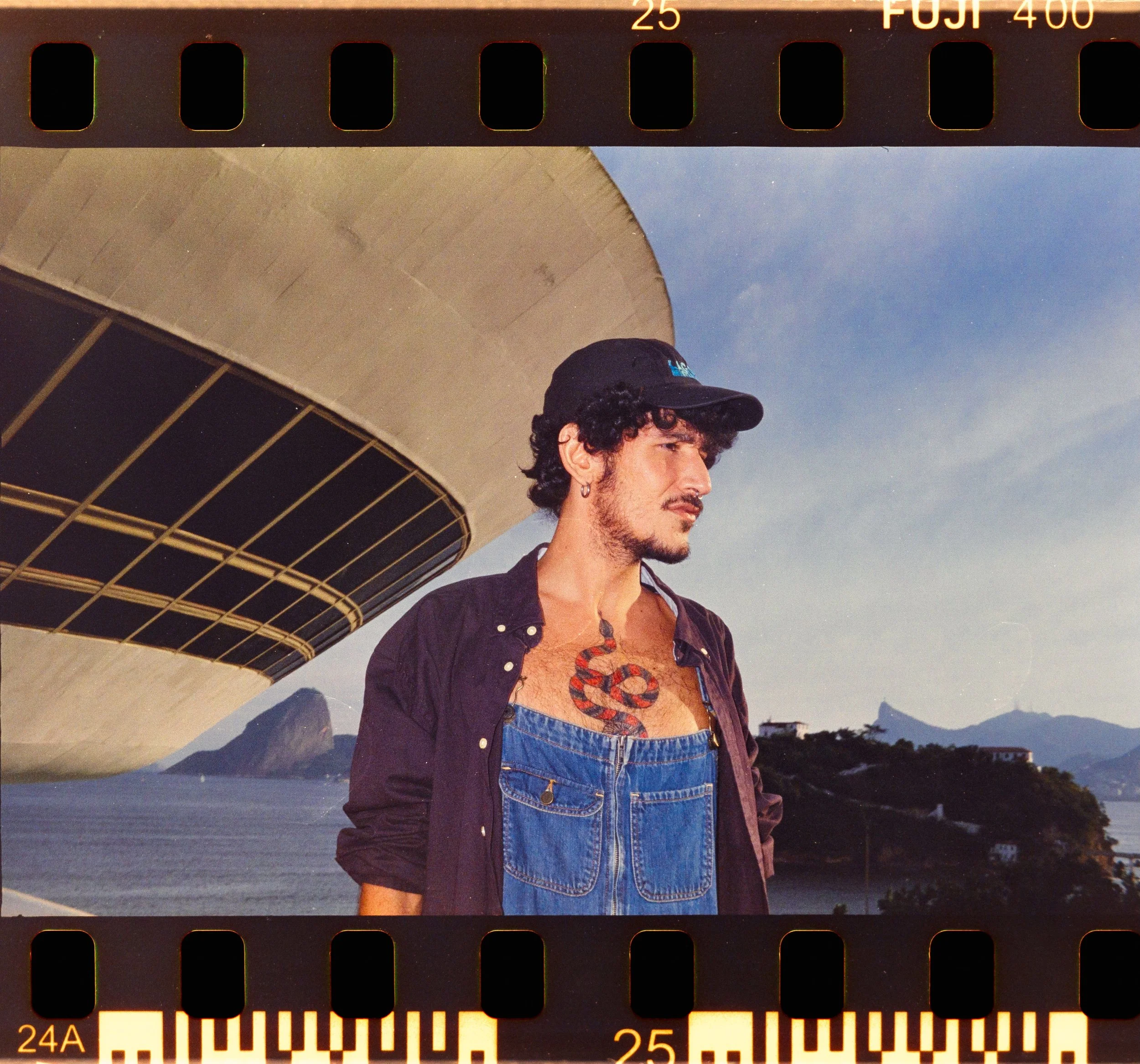 A man with curly dark hair, wearing a black cap, earrings, a dark shirt, and denim overalls, standing outdoors near a large, modern building with a curved design, in a coastal area with hills and water in the background.