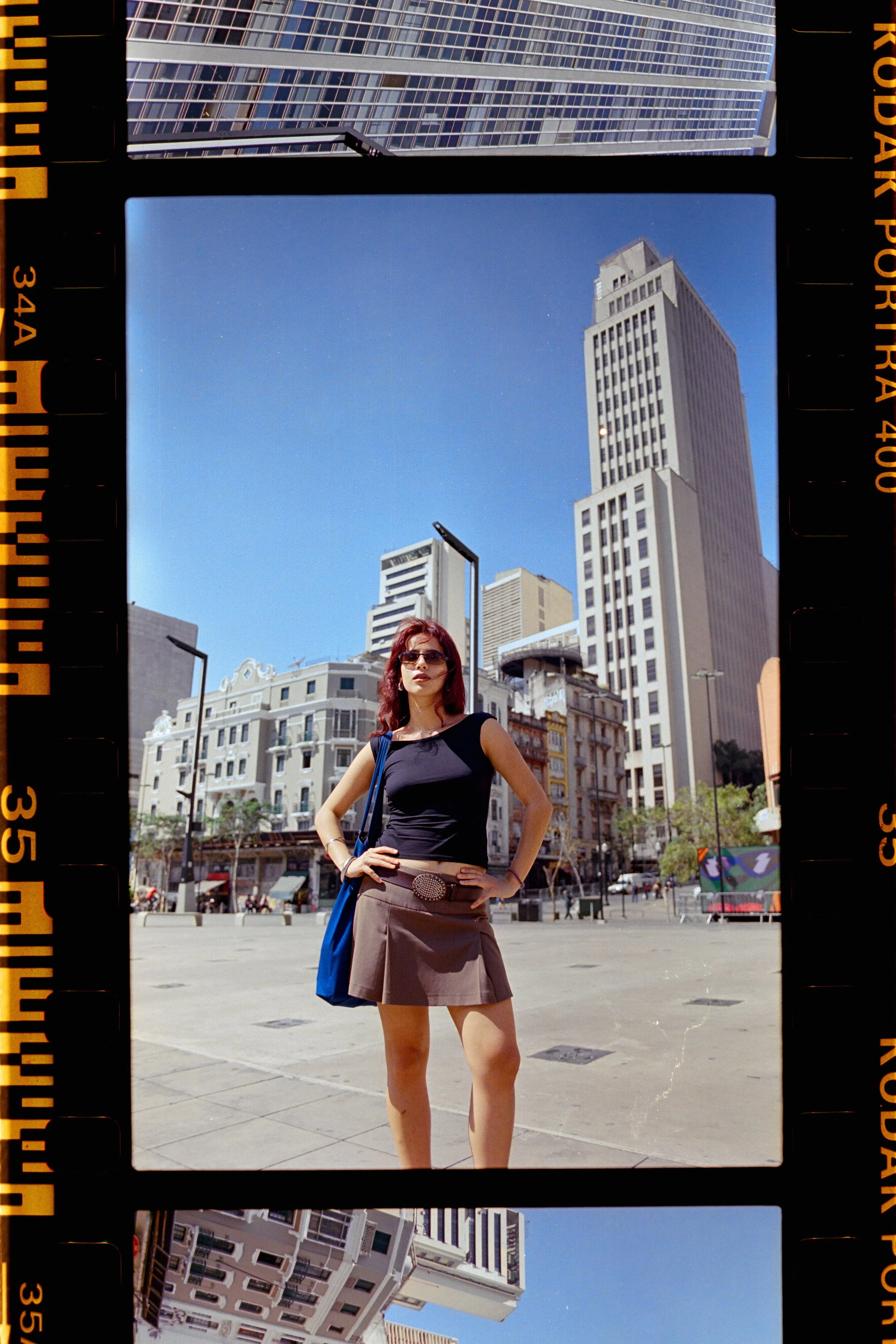 A woman with red hair wearing sunglasses, a black sleeveless top, a brown skirt with a belt, and a blue bag stands under a clear blue sky in an urban area with tall buildings and historic architecture, framed by black and orange film strip borders.
