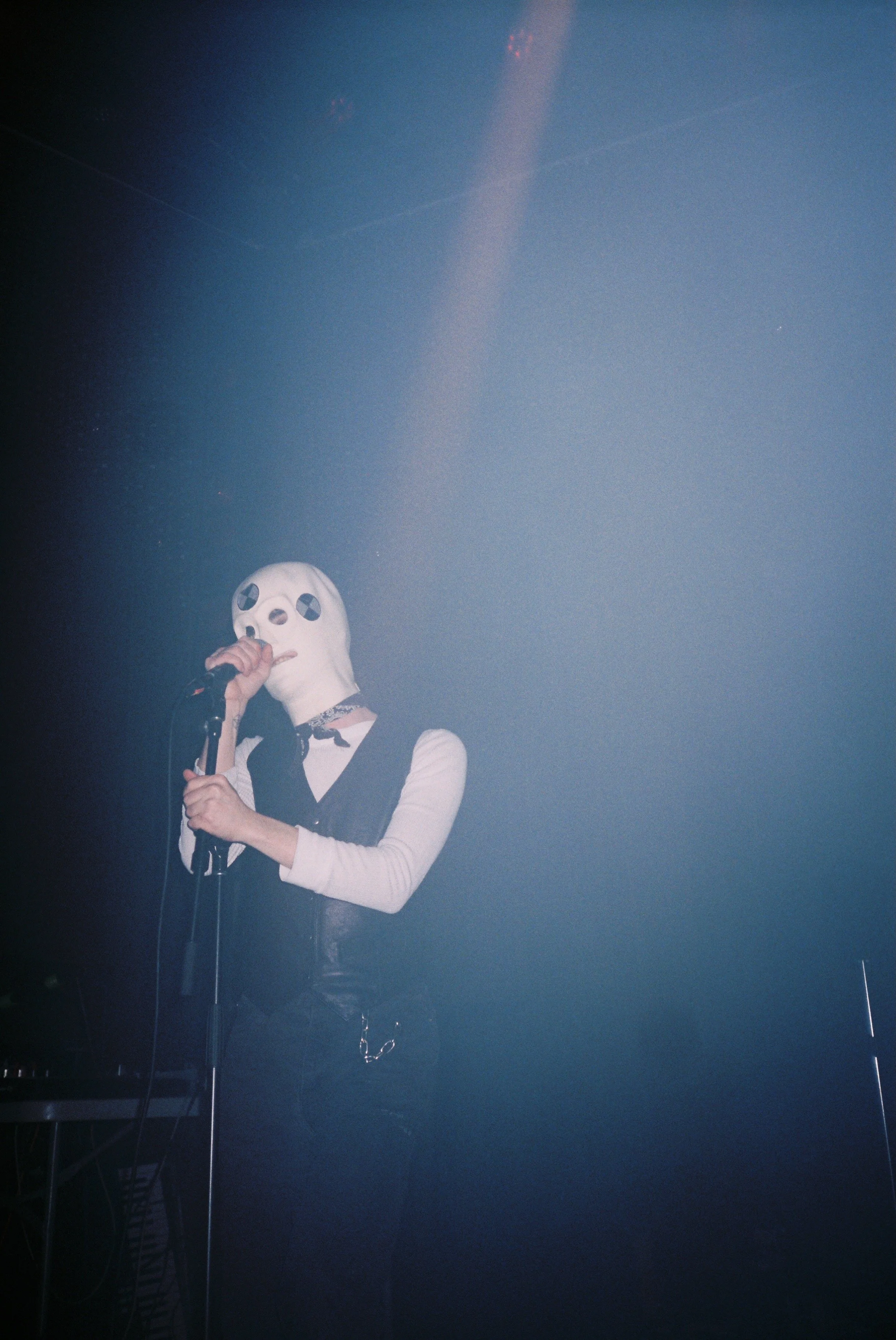 A person wearing a white mask with black eyes and a mouth, holding a microphone, dressed in a black vest over a white long-sleeve shirt, standing on stage with fog and blue lighting, performing.