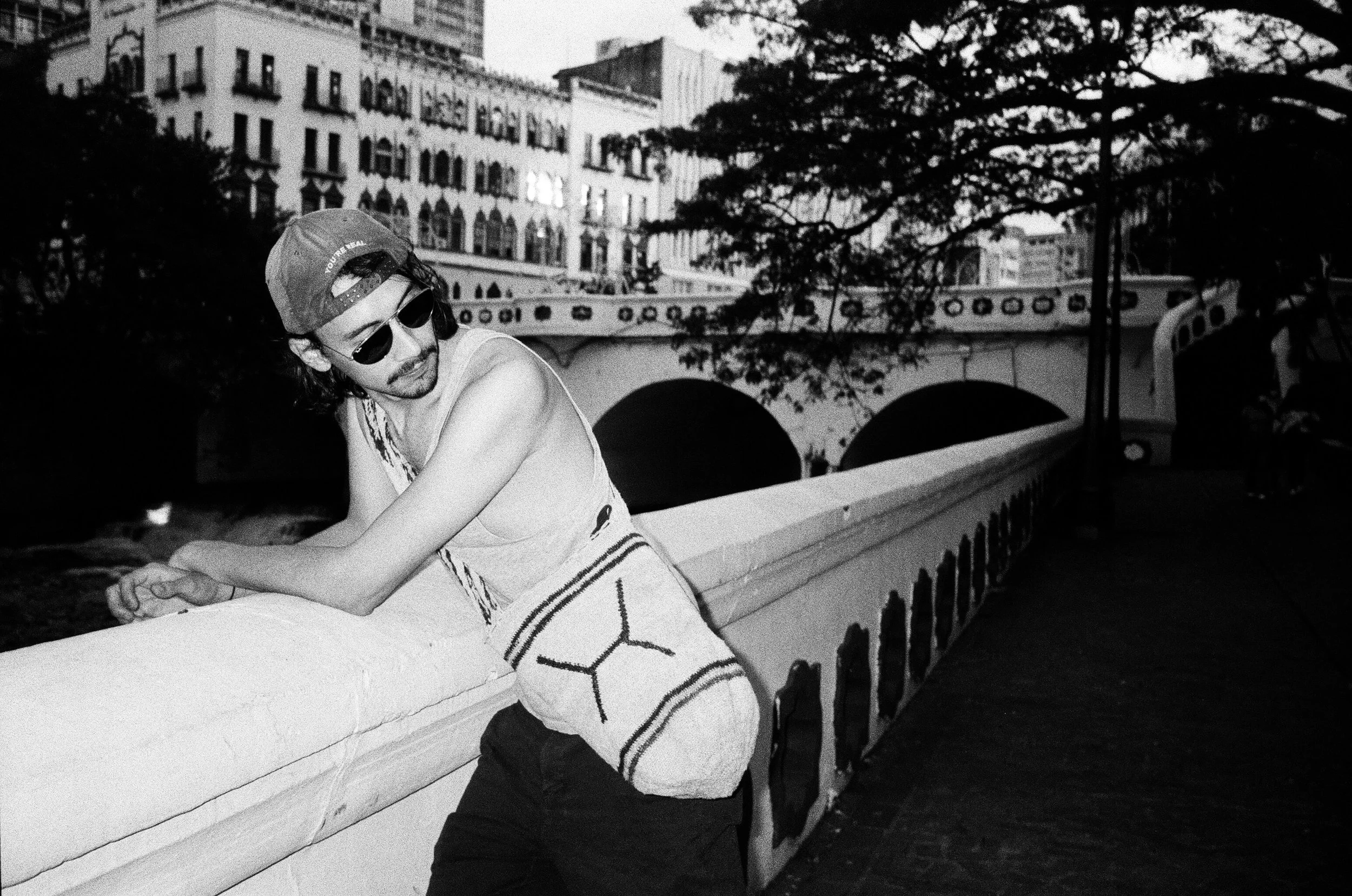 A man leaning on a white railing outdoors during daytime, wearing sunglasses, a cap backward, and a tank top, with buildings and trees in the background.