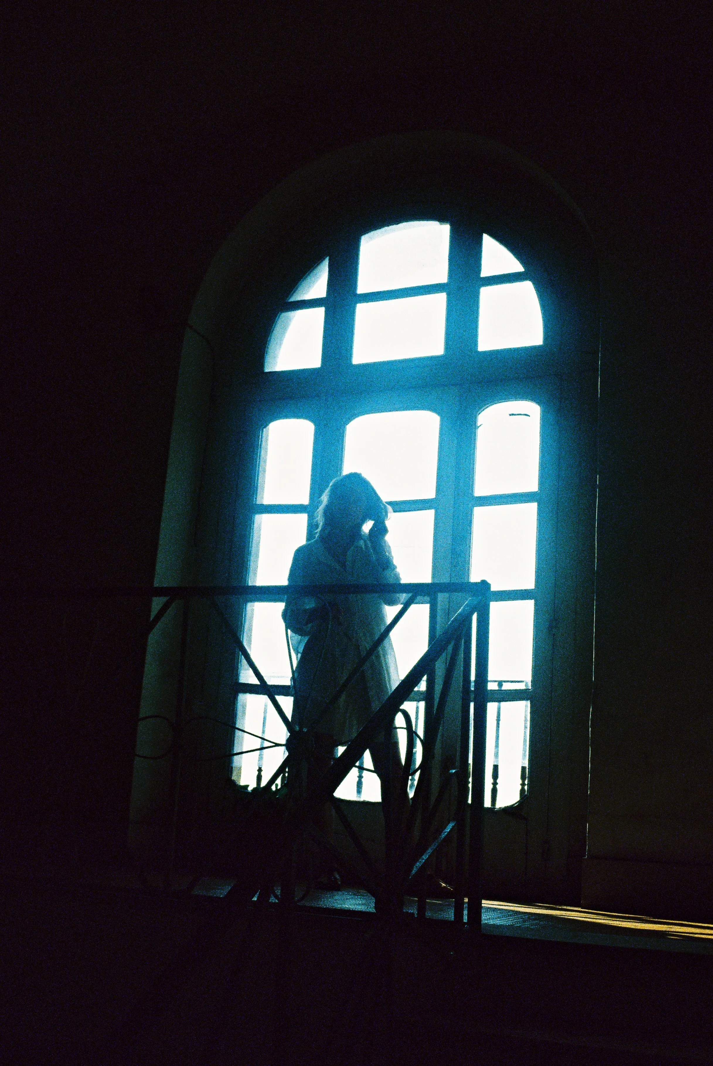 A person standing on a balcony inside a building, talking on a cell phone, with a large arched window behind them that lets in bright light, creating a silhouette effect.