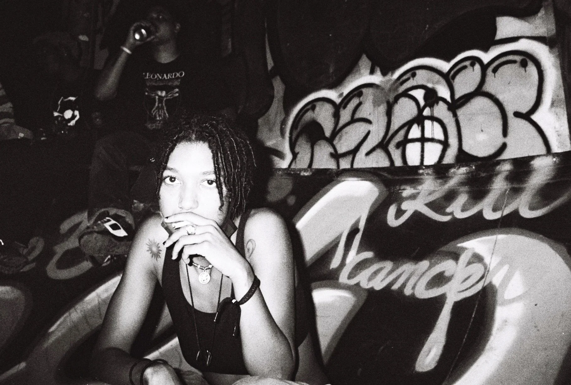A young woman with dreadlocks covering her forehead, wearing a sleeveless top, sitting in front of a graffiti-covered wall with large, stylized lettering. She has multiple necklaces, a tattoo near her shoulder, and is holding her hand to her mouth, gazing directly at the camera.