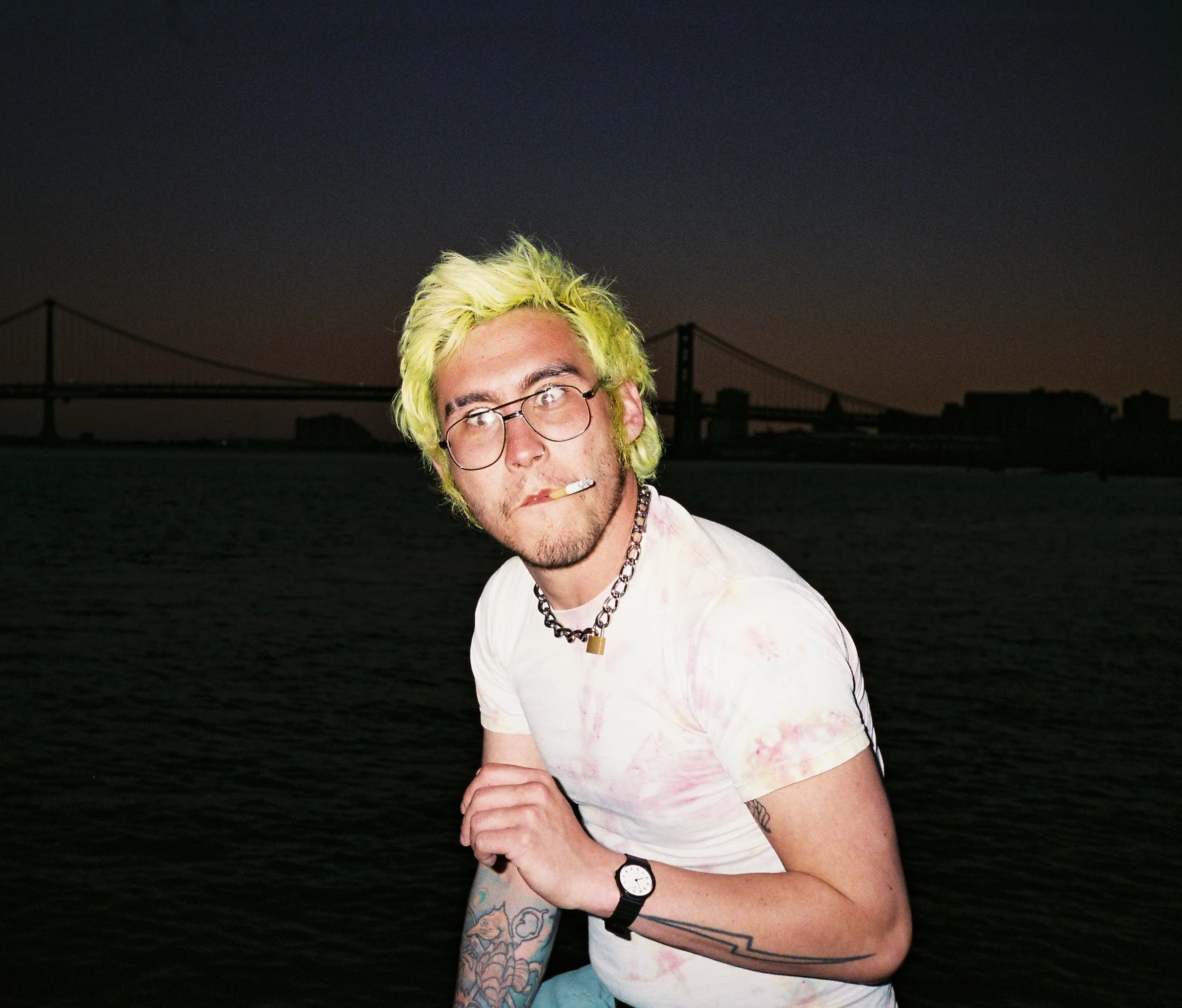A man with yellow-green hair, glasses, and tattoos on his arm, wearing a white tie-dye shirt, standing outdoors at night by the water with a bridge in the background.