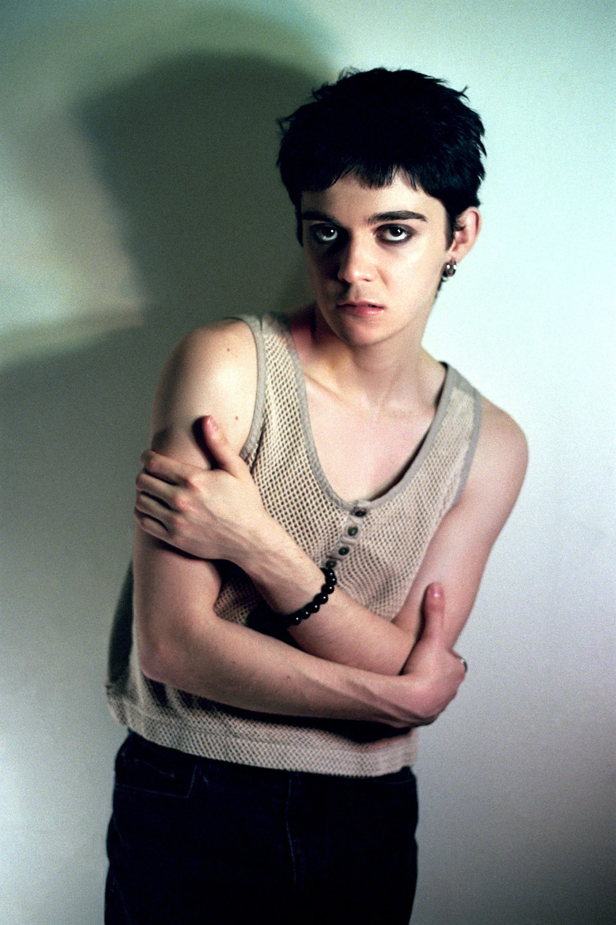 A young woman with short dark hair and dark eye makeup posing against a plain wall, wearing a sleeveless beige mesh top and black pants, with her arms crossed over her chest.