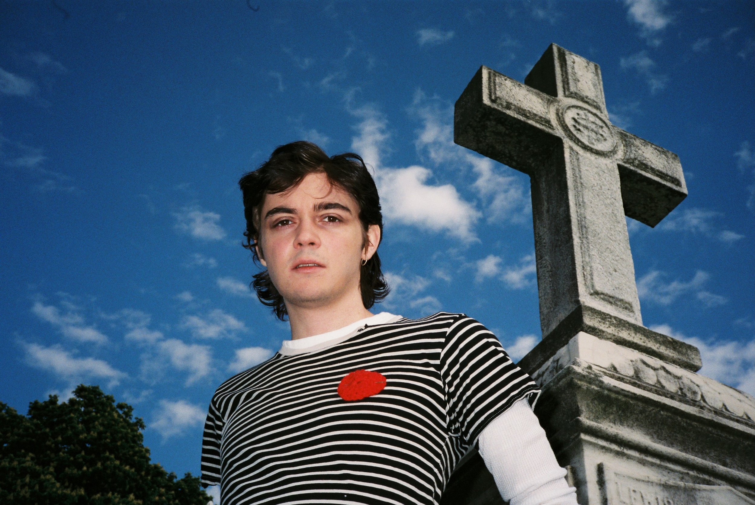 A young man with dark hair and an earring, wearing a black and white striped shirt with a red poppy pin, stands outdoors near a large stone cross monument on a bright day with blue skies and white clouds in the background.