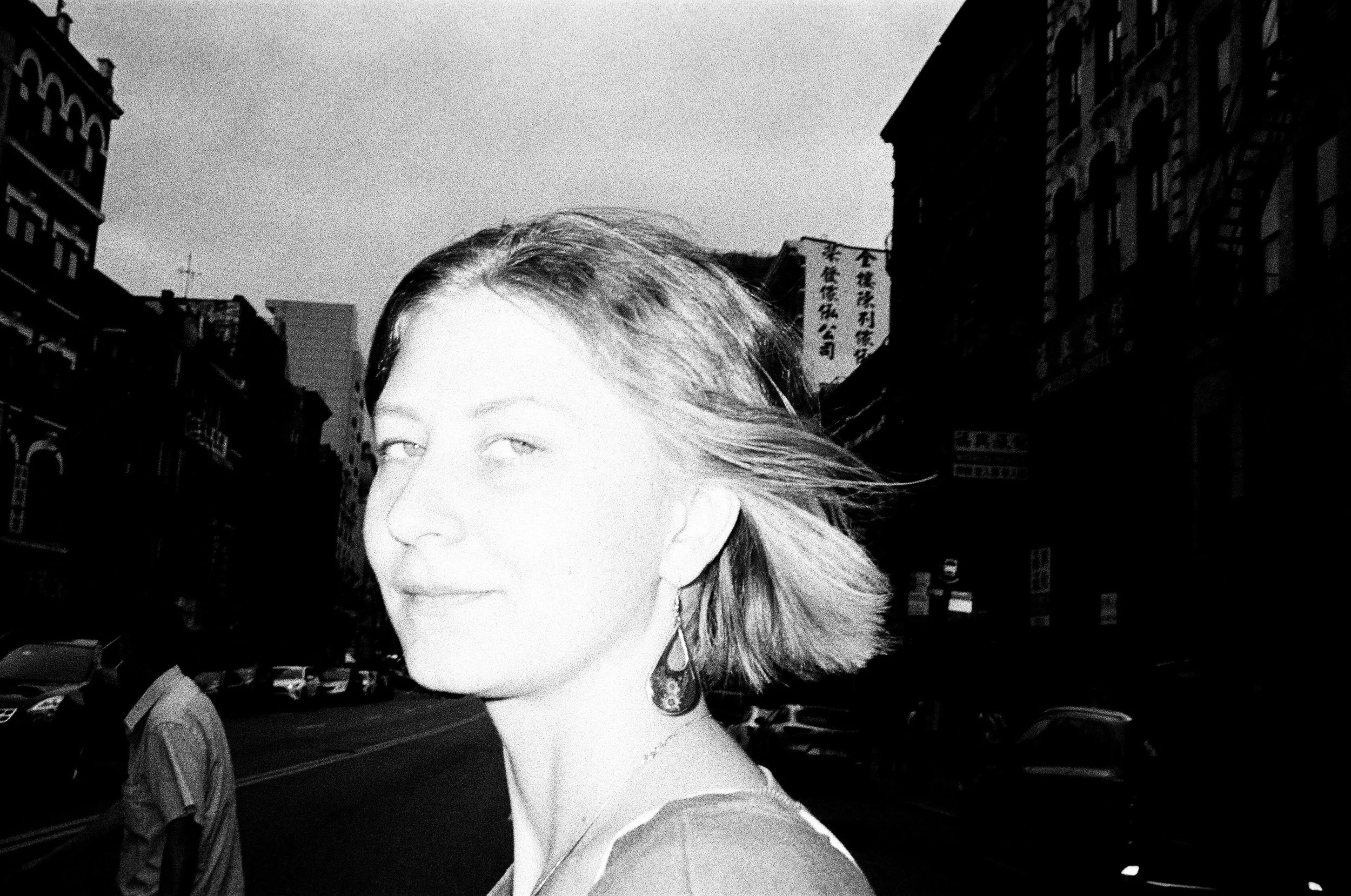 Black and white photo of a woman with short hair on an urban street, with buildings and parked cars in the background.
