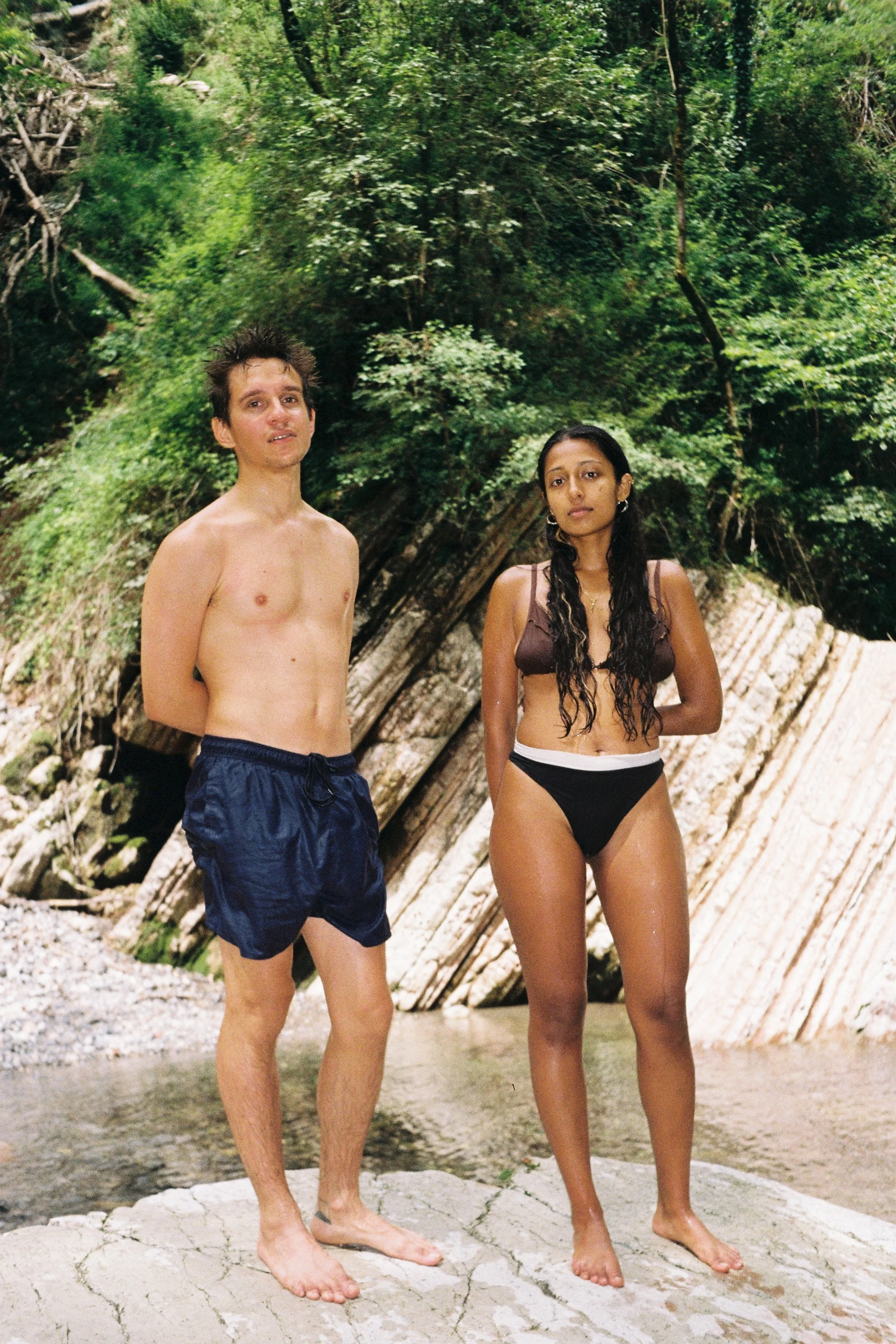 A man and woman standing barefoot on a rock by a river with lush greenery and large rock formations in the background.