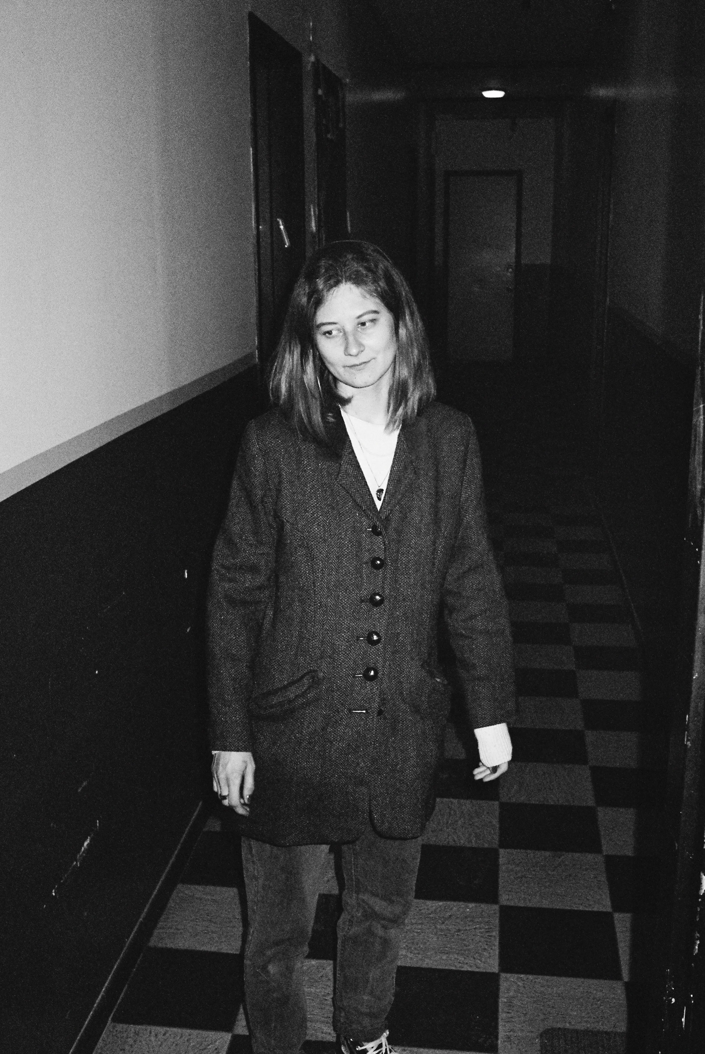 A young woman with shoulder-length hair standing in a hallway with a checkered floor, wearing a blazer over a white shirt and pants, in black and white photo.