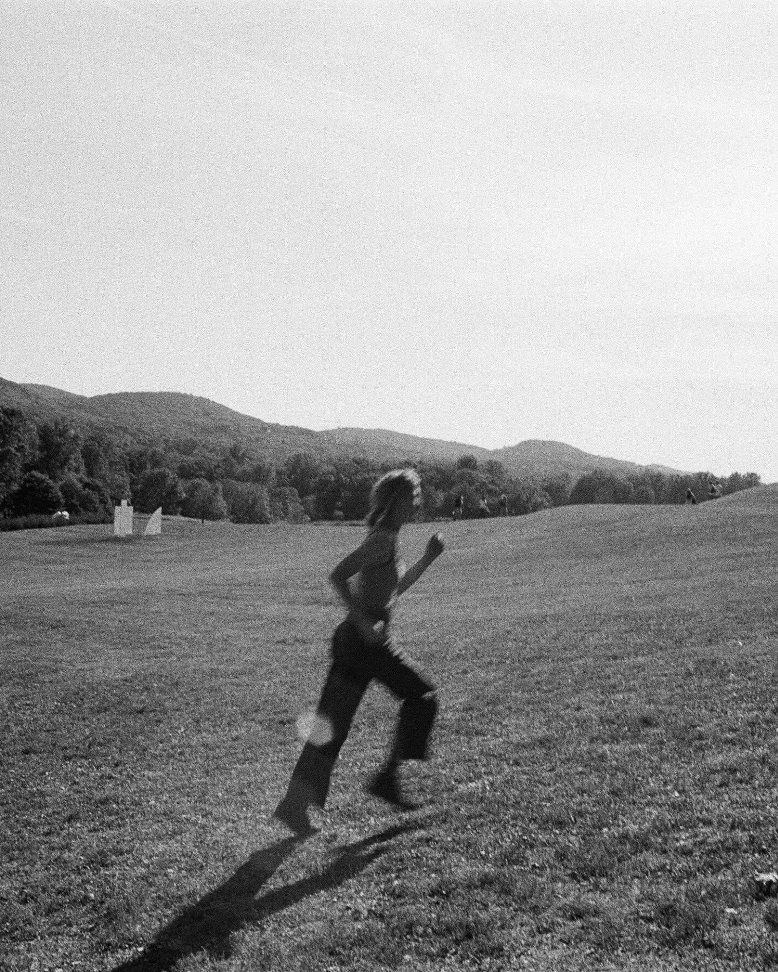 A person running across a grassy field with rolling hills and trees in the background, in black and white.