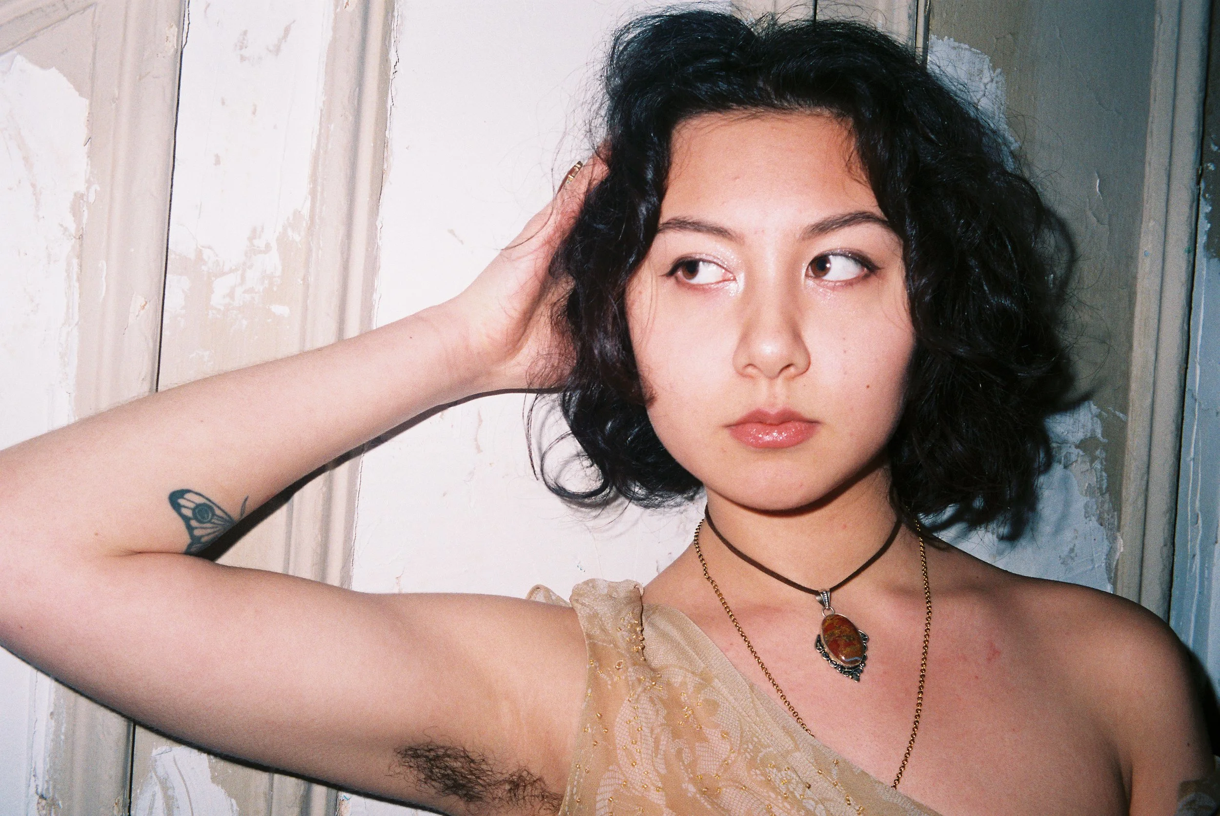 A young woman with black curly hair and light skin, wearing a beige sleeveless top and layered necklaces, standing against a distressed white wooden wall, looking to her right with her left hand touching her hair.