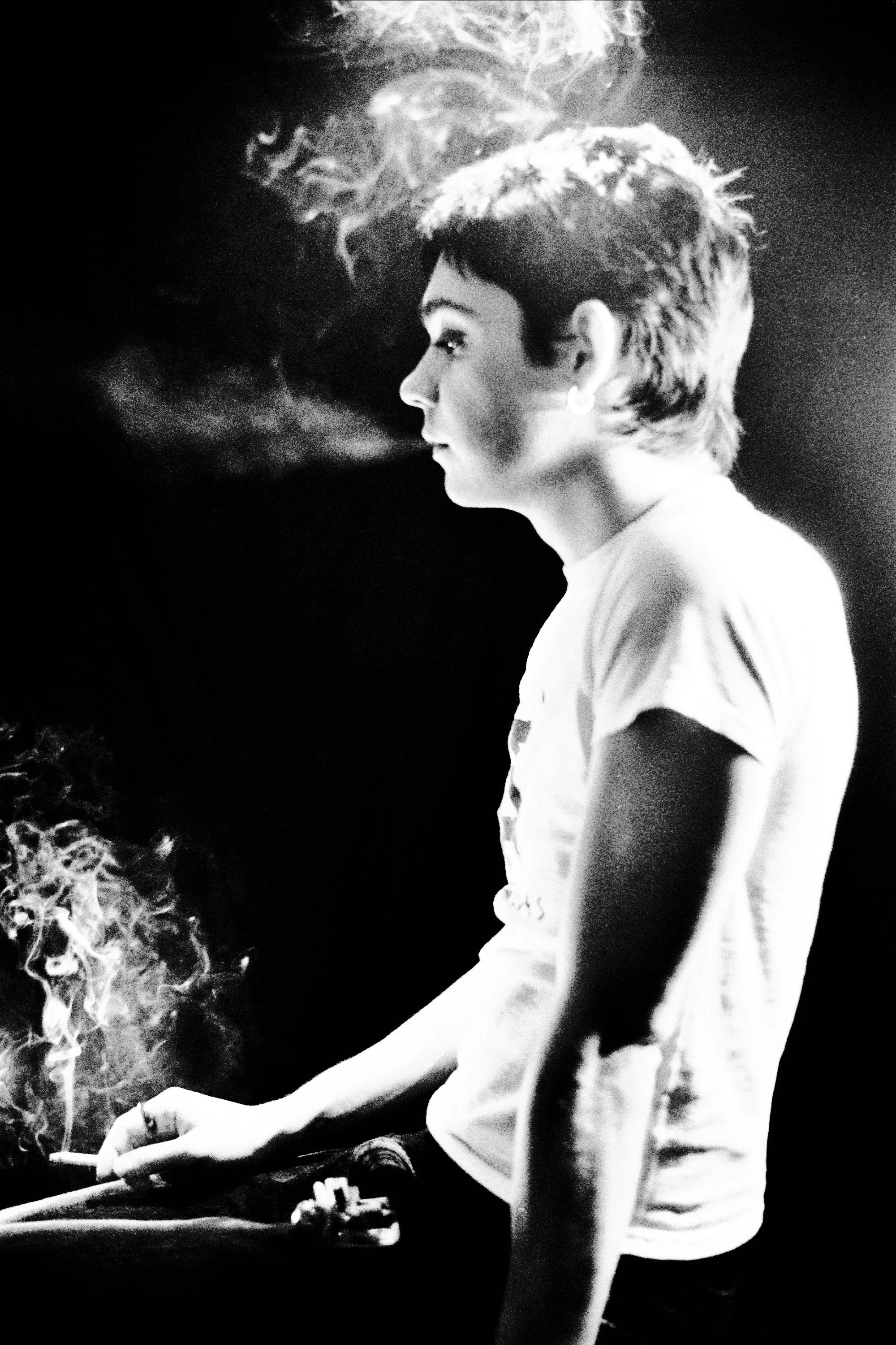 Black and white photo of a young woman with short hair, profile view, smoking a cigarette with smoke swirling around her head.