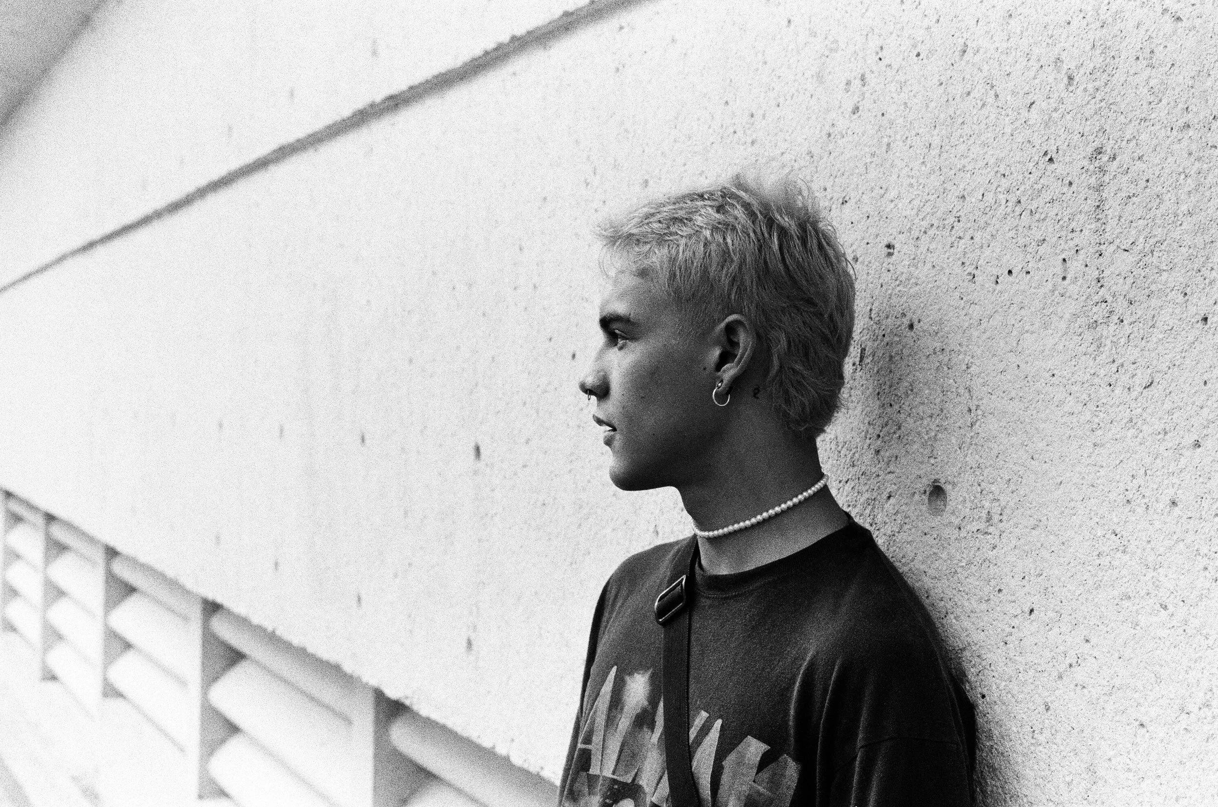 A young person with short, light-colored hair and earrings, wearing a pearl necklace and a dark graphic T-shirt, stands against a textured concrete wall, looking to the side.