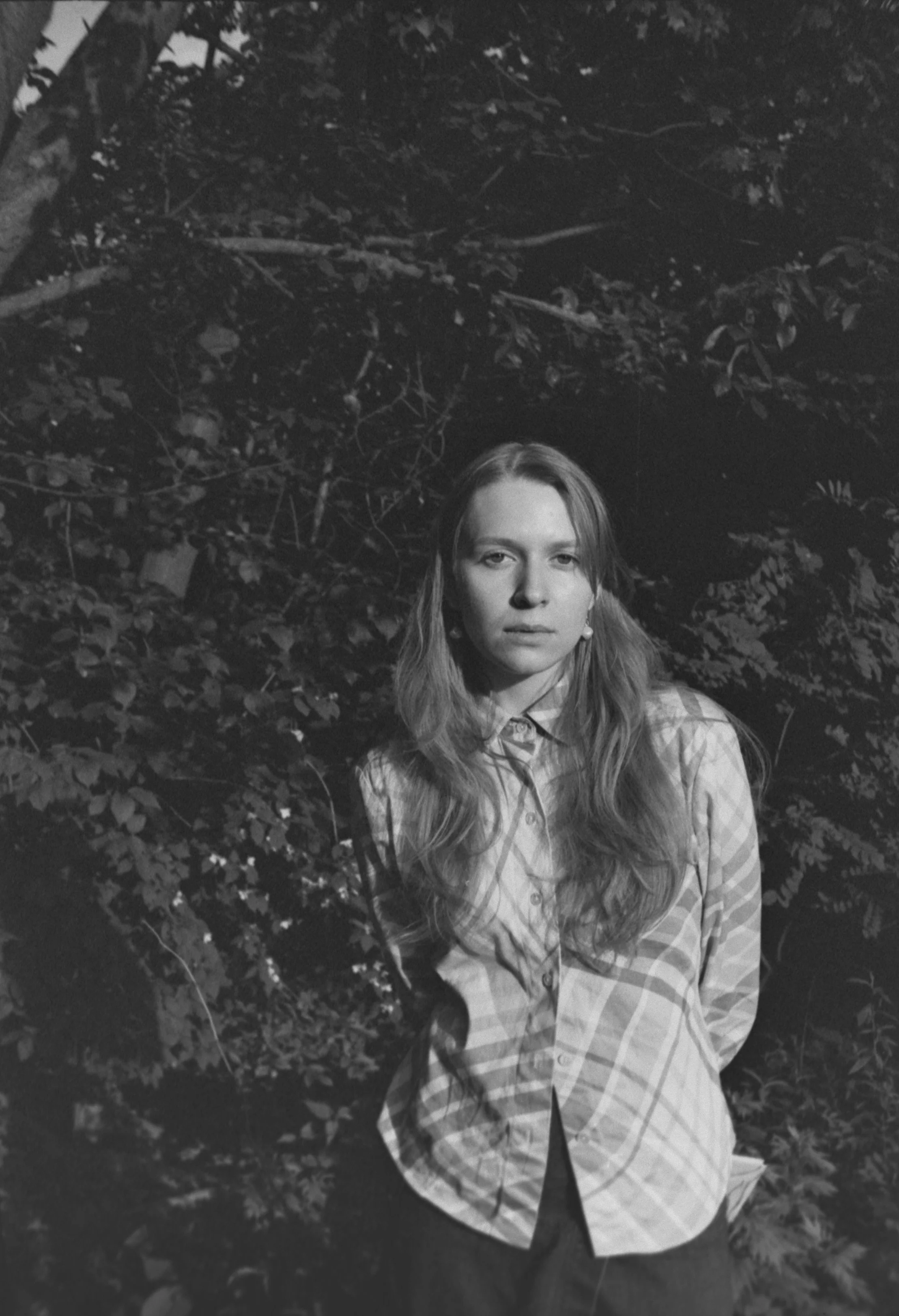A black and white photo of a young woman with long hair, standing in front of a dense background of leaves and branches. She is wearing a plaid button-up shirt with hands behind her back and looking directly at the camera.