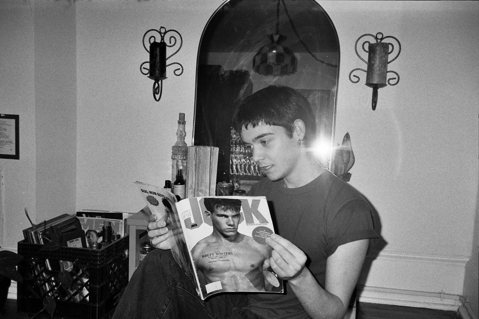 A young person with short dark hair, wearing a short-sleeved shirt, is sitting and reading a magazine featuring a male model with muscular build on the cover, in a room with a mirror, wall-mounted candle sconces, and various bottles and magazines on 