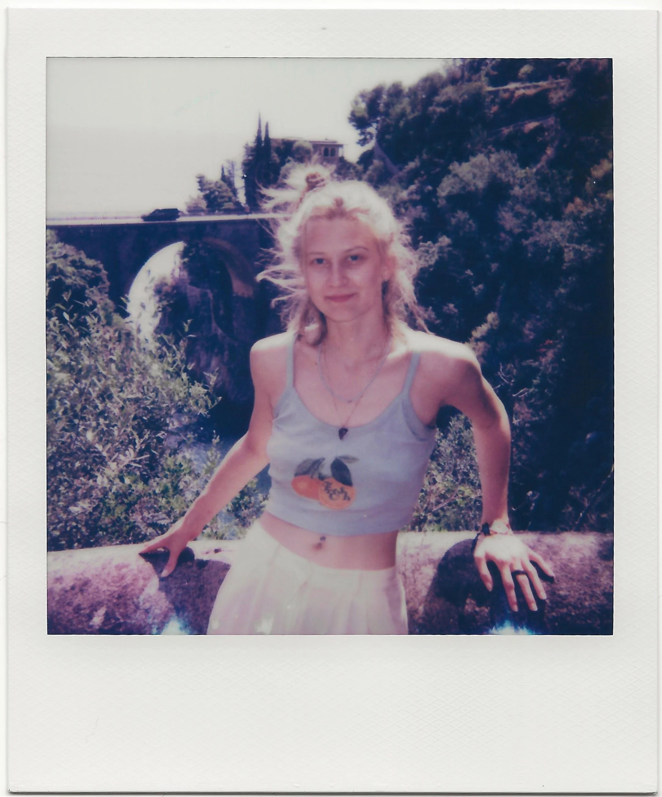A young woman with blonde hair standing outdoors, leaning on a stone ledge with greenery and trees in the background, and a bridge or aqueduct visible in the distance.