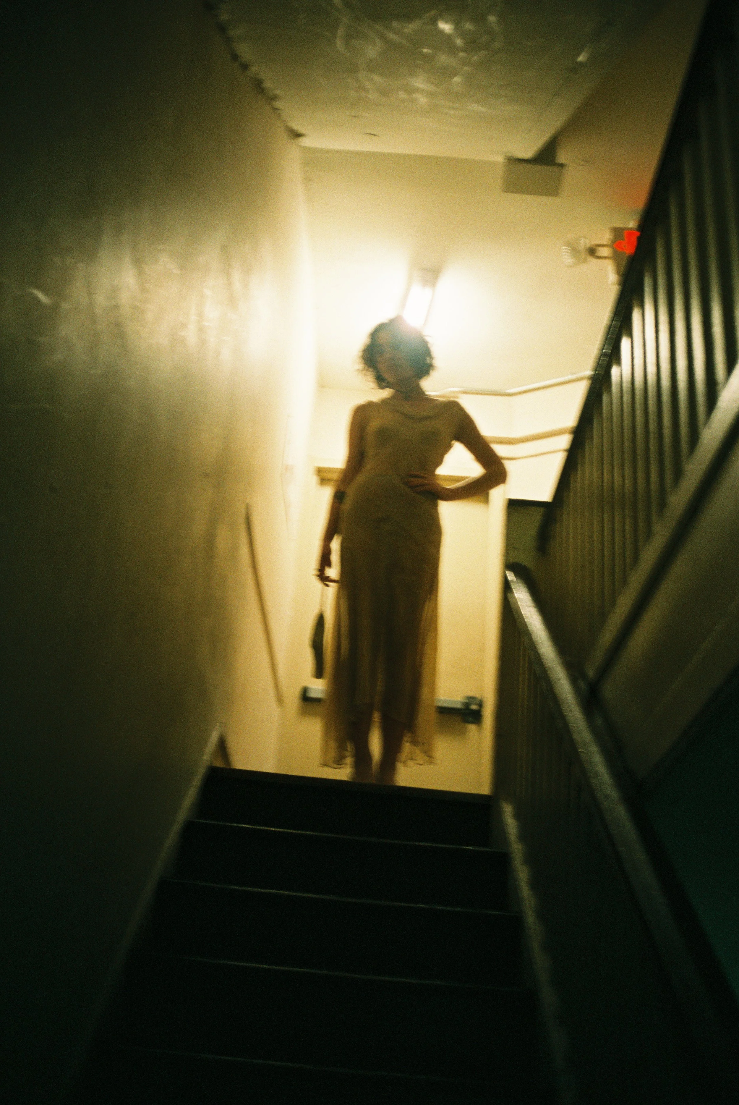 A woman standing at the top of a staircase, backlit by a bright light overhead, with one hand on her hip and holding a purse in the other, wearing a sheer dress.