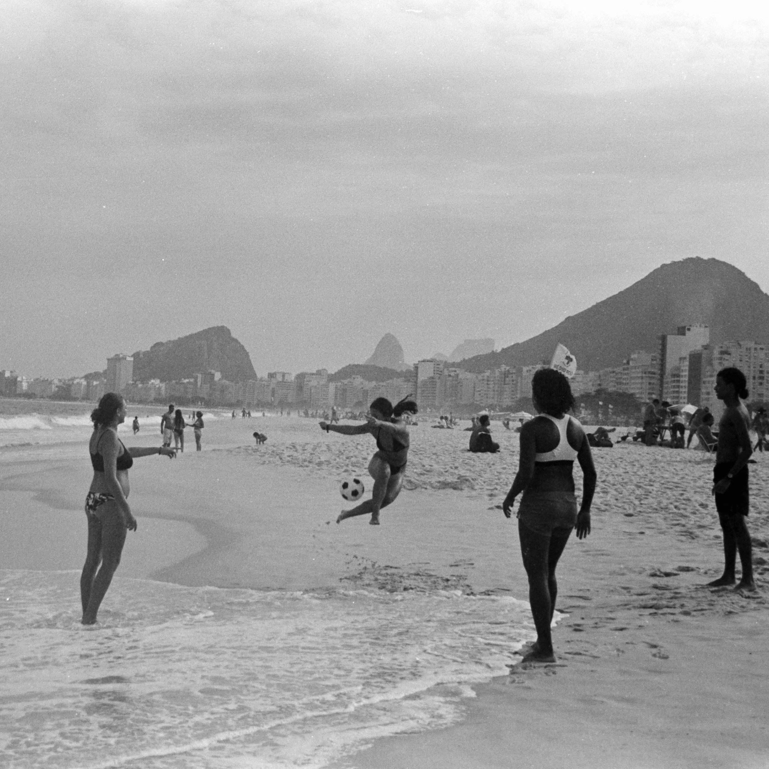 People playing soccer on a beach with a city skyline and mountains in the background.