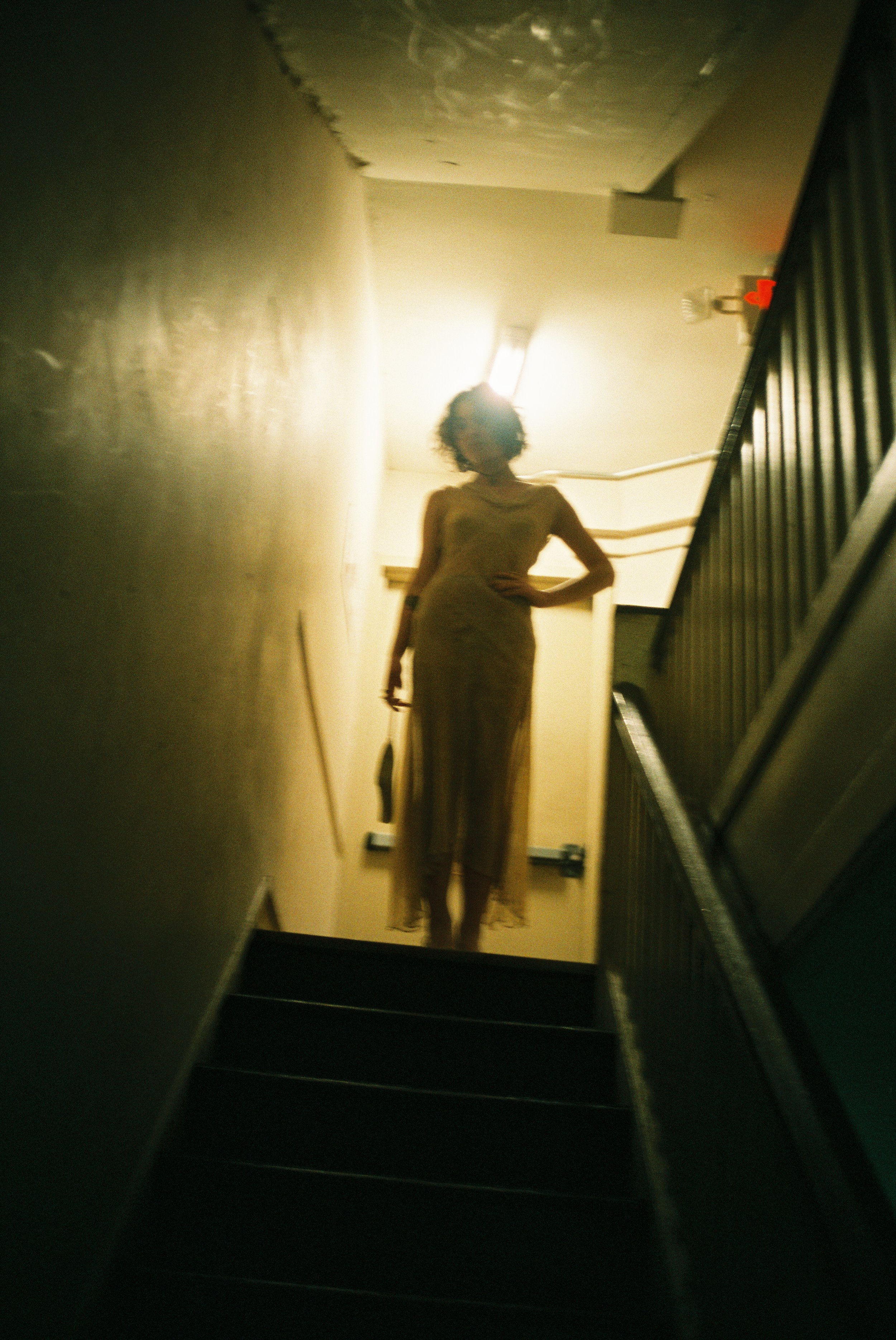 A woman with curly hair standing at the top of a staircase, backlit by a bright ceiling light, wearing a long dress and holding an object in her left hand.