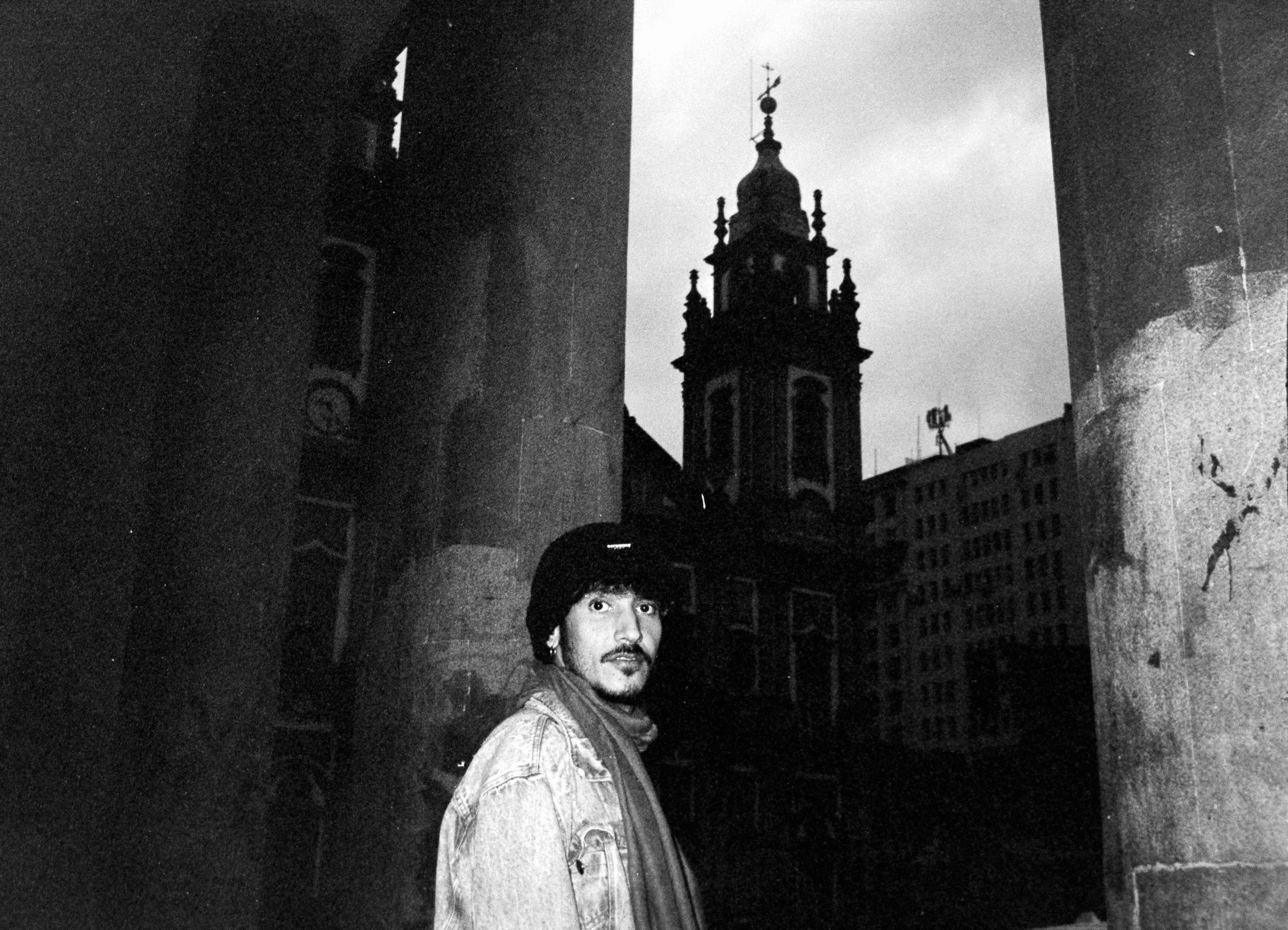 A man with dark hair and a beard wearing a denim jacket and scarf, standing outdoors with large stone columns on either side, a church with a tall steeple in the background, and the sky visible.