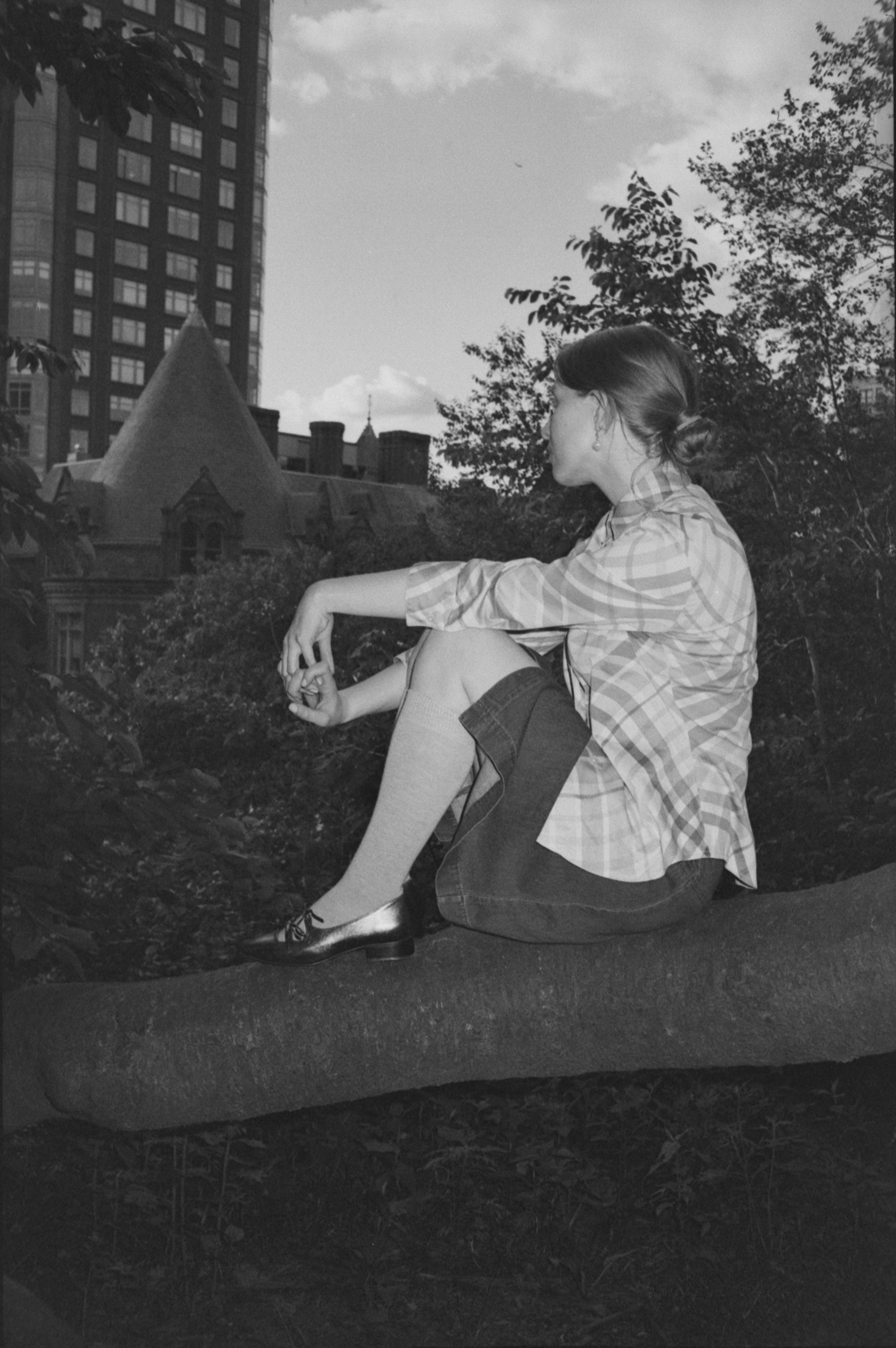A young woman with patterned shirt, knee-high socks, and shiny shoes sitting on a tree branch in a city park during daytime, with buildings and trees in the background.