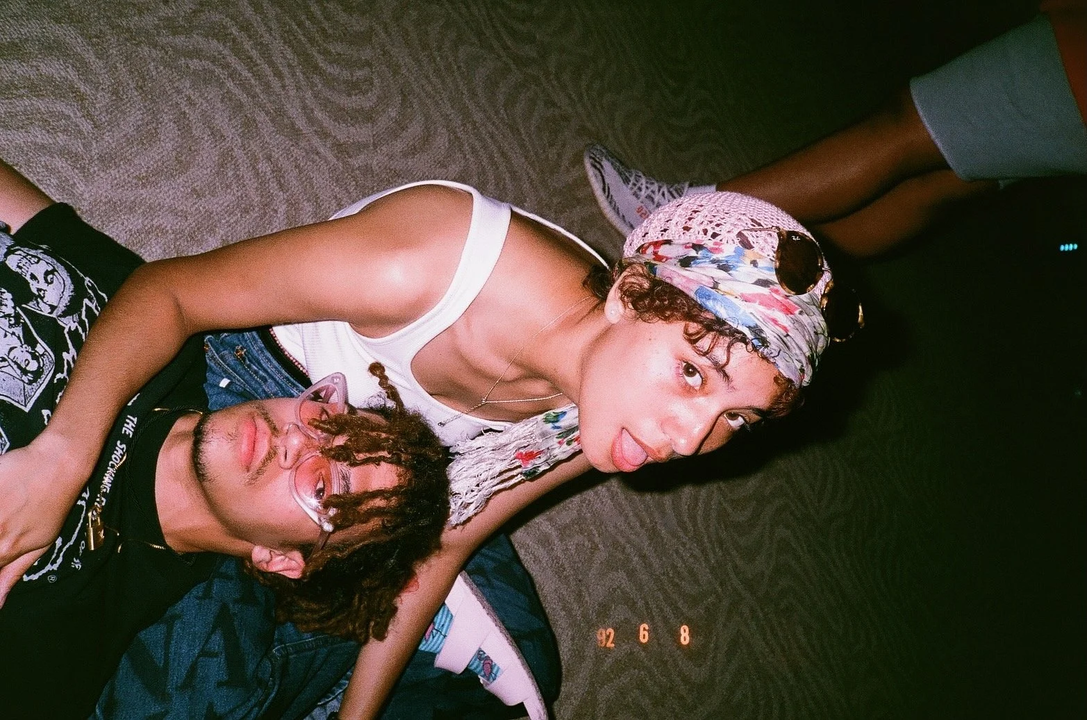 A woman with curly hair and sunglasses on her head, lying on the floor, sticking out her tongue, with a person with dreadlocks and glasses resting their head on her arm.