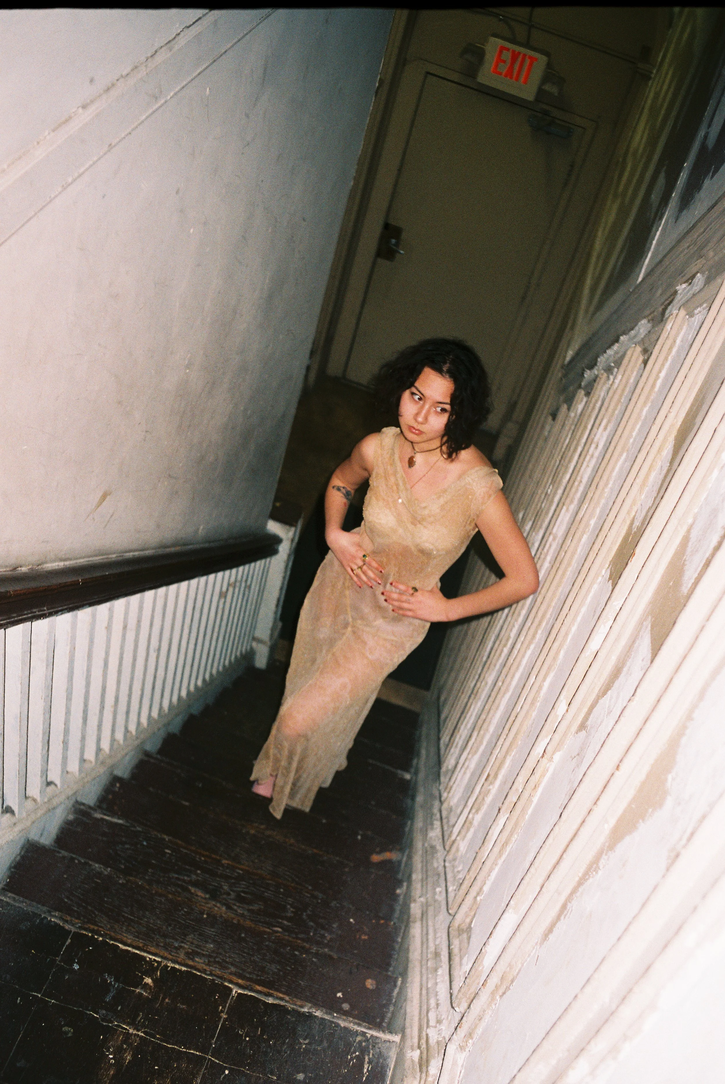 A woman with dark, curly hair wearing a beige, semi-transparent dress stands on a narrow wooden staircase with her hands on her hips. She is near a wall with peeling paint and a staircase railing. There is an exit sign above a door at the top of the 