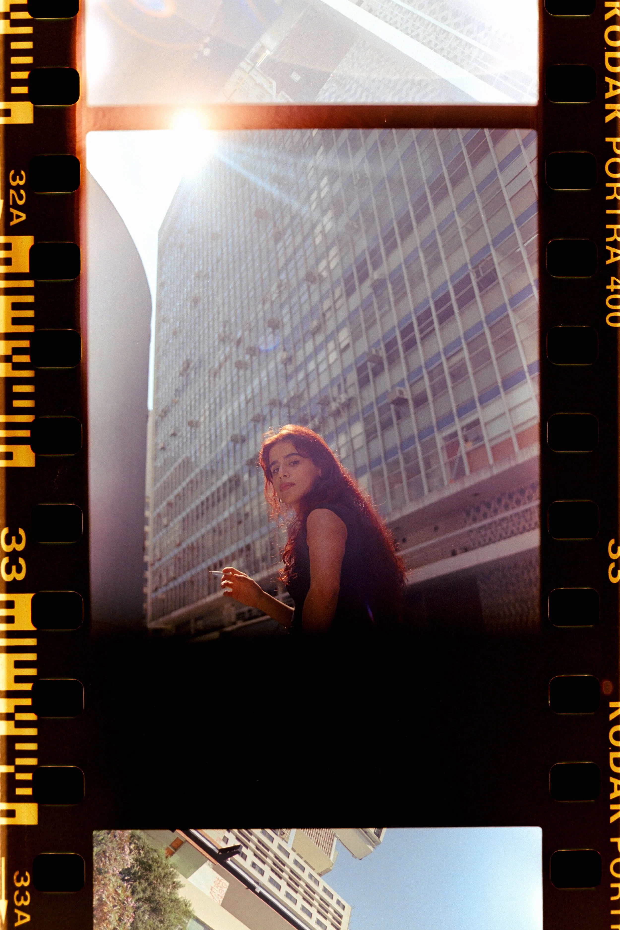 A woman with long, dark hair standing outdoors in front of a modern glass building, with bright sunlight overhead.