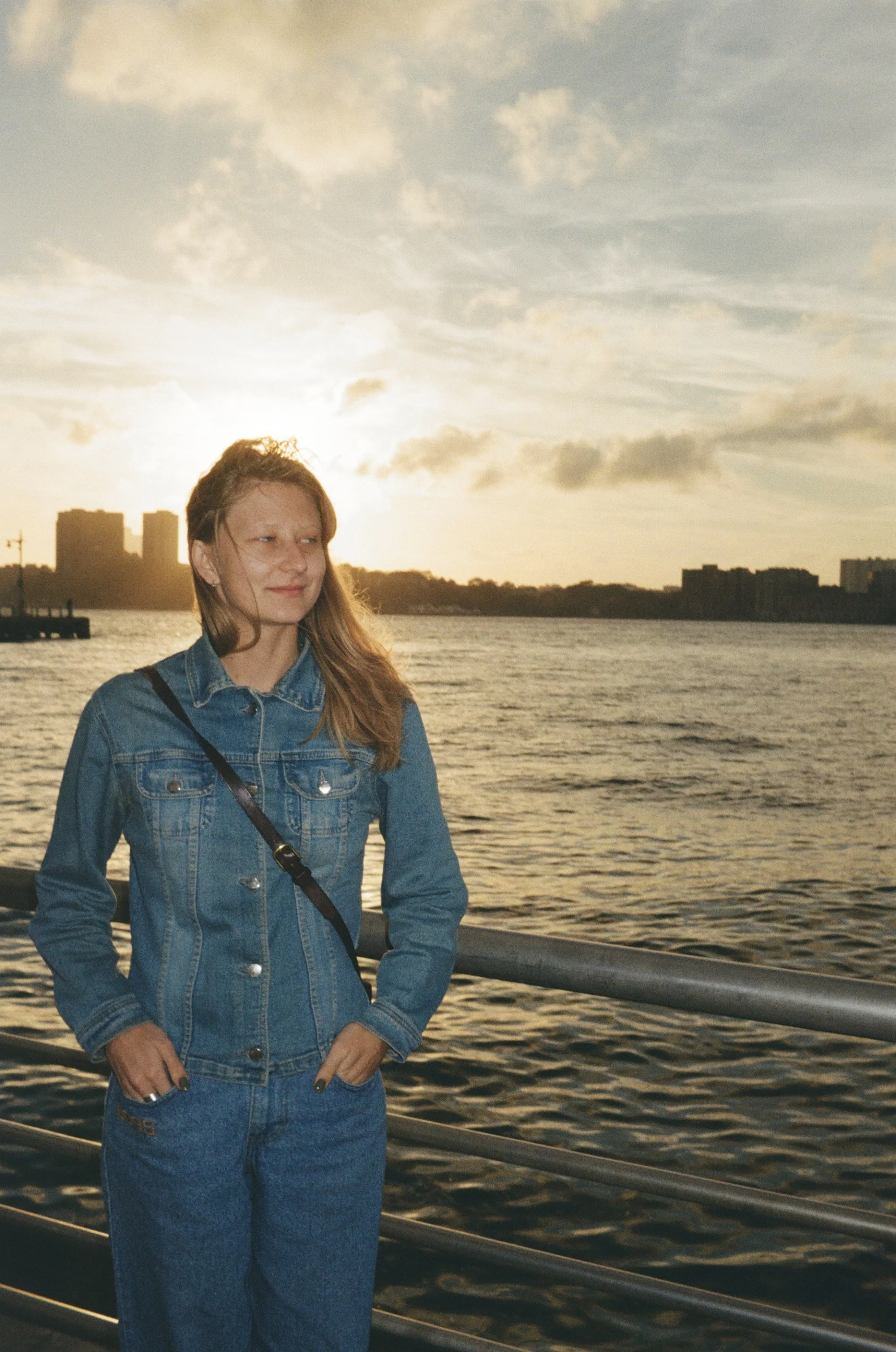 A woman in a denim jacket and jeans standing by a body of water during sunset.
