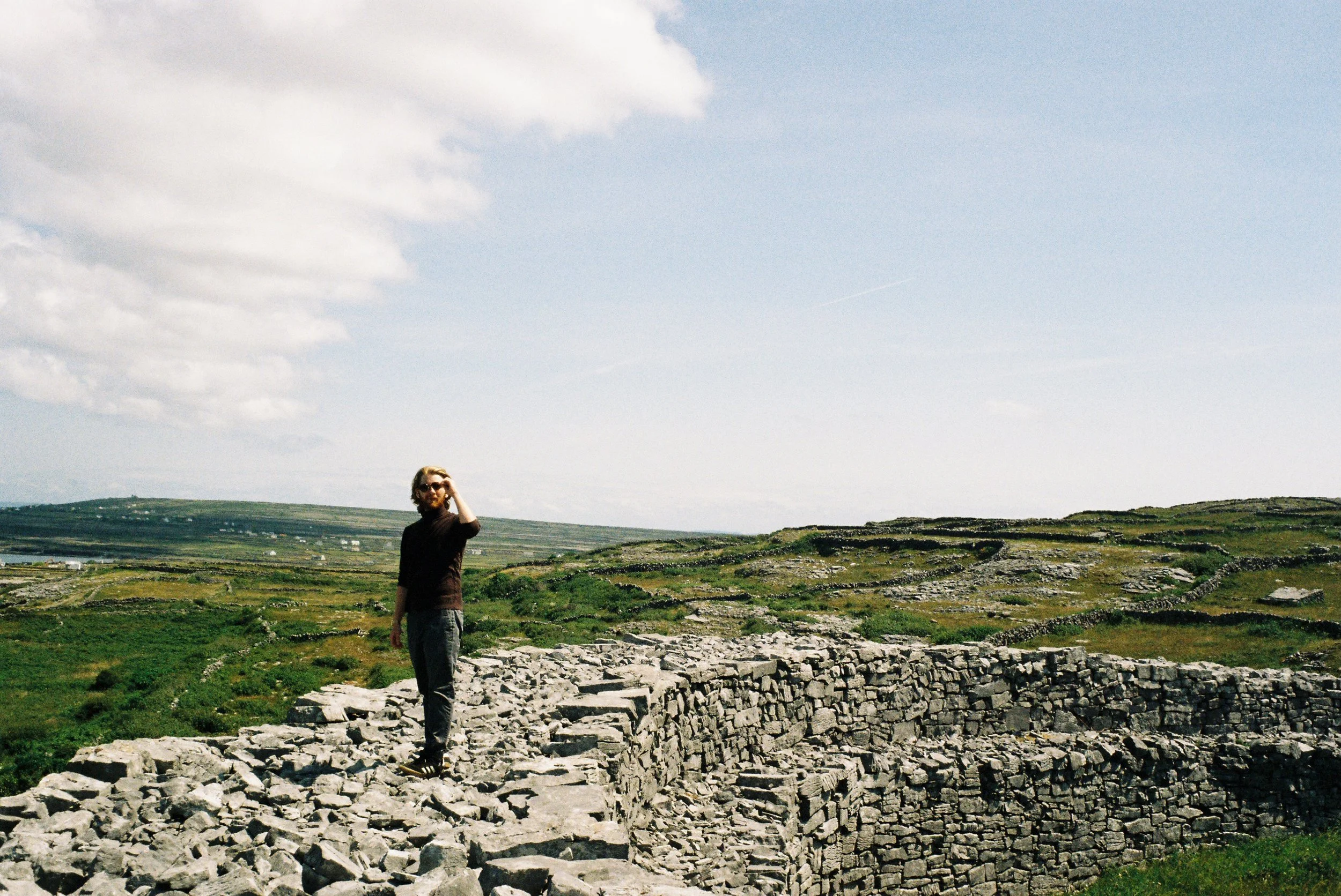 A person standing on ancient stone ruins in a grassy landscape under a partly cloudy sky.