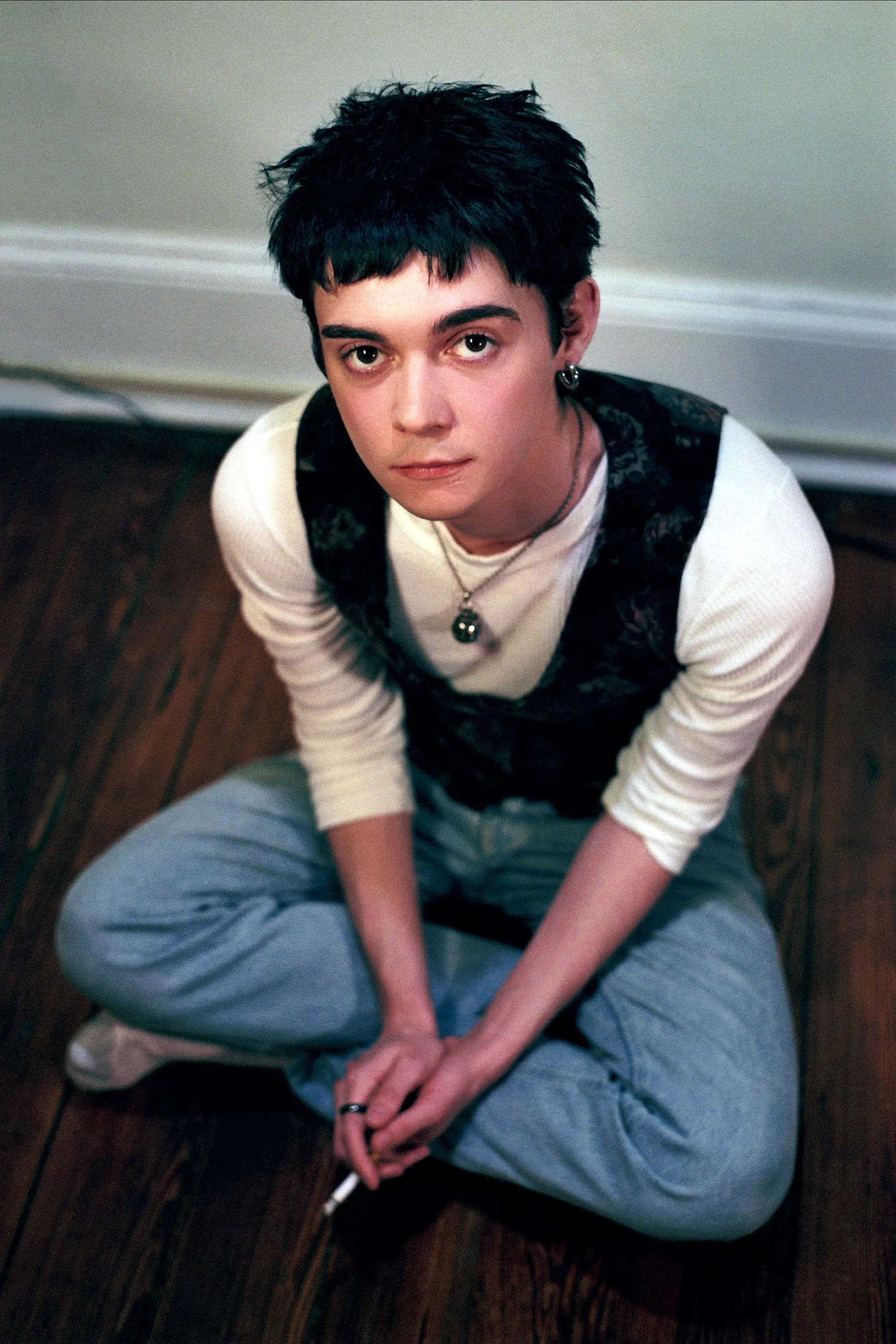 Young person with short dark hair and multiple earrings, sitting on a wooden floor with legs crossed, looking up at the camera, holding a cigarette in their hands.