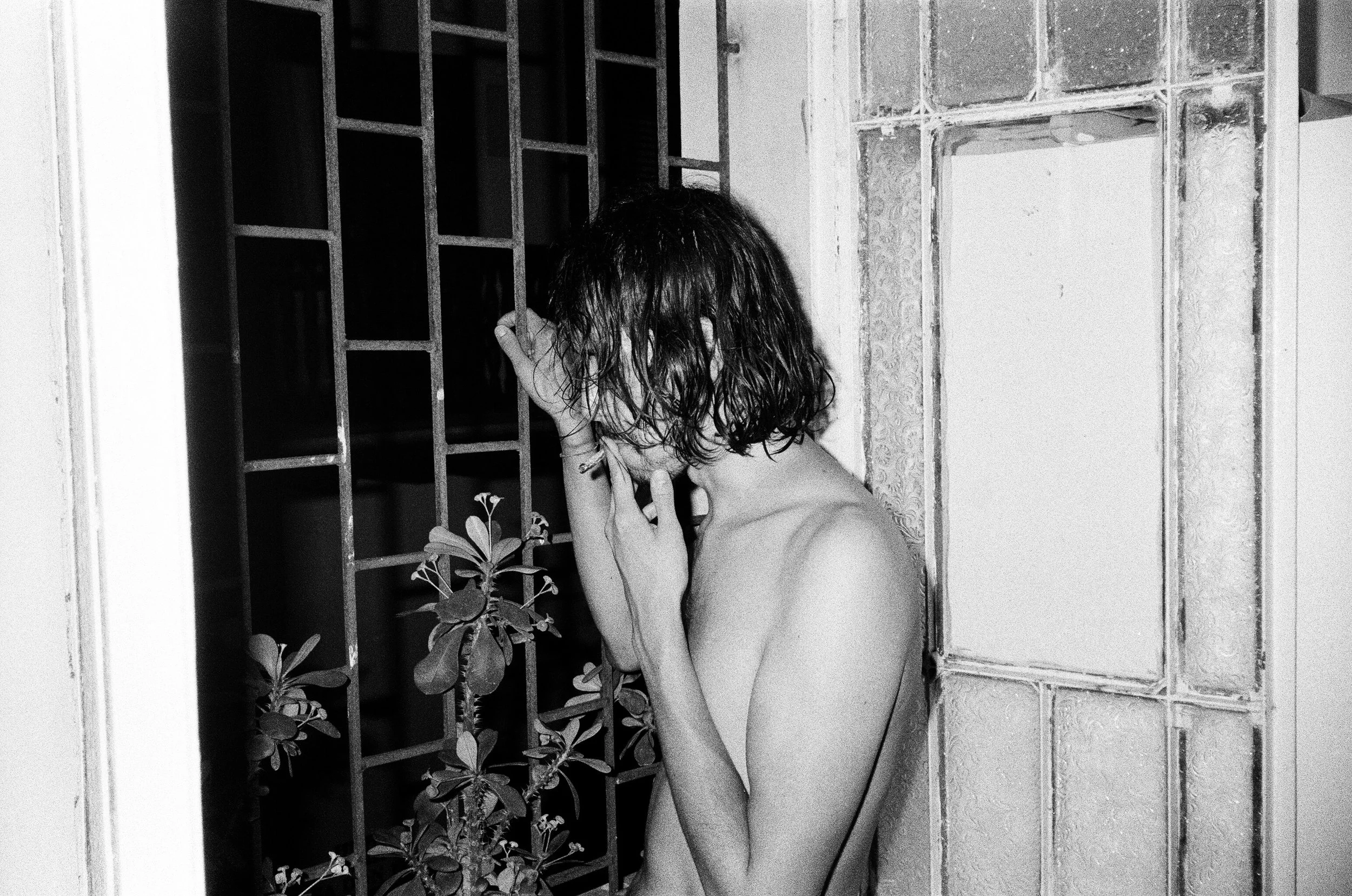 A topless woman with wet hair stands behind a metal gate, holding her face and looking down in a dimly lit area.
