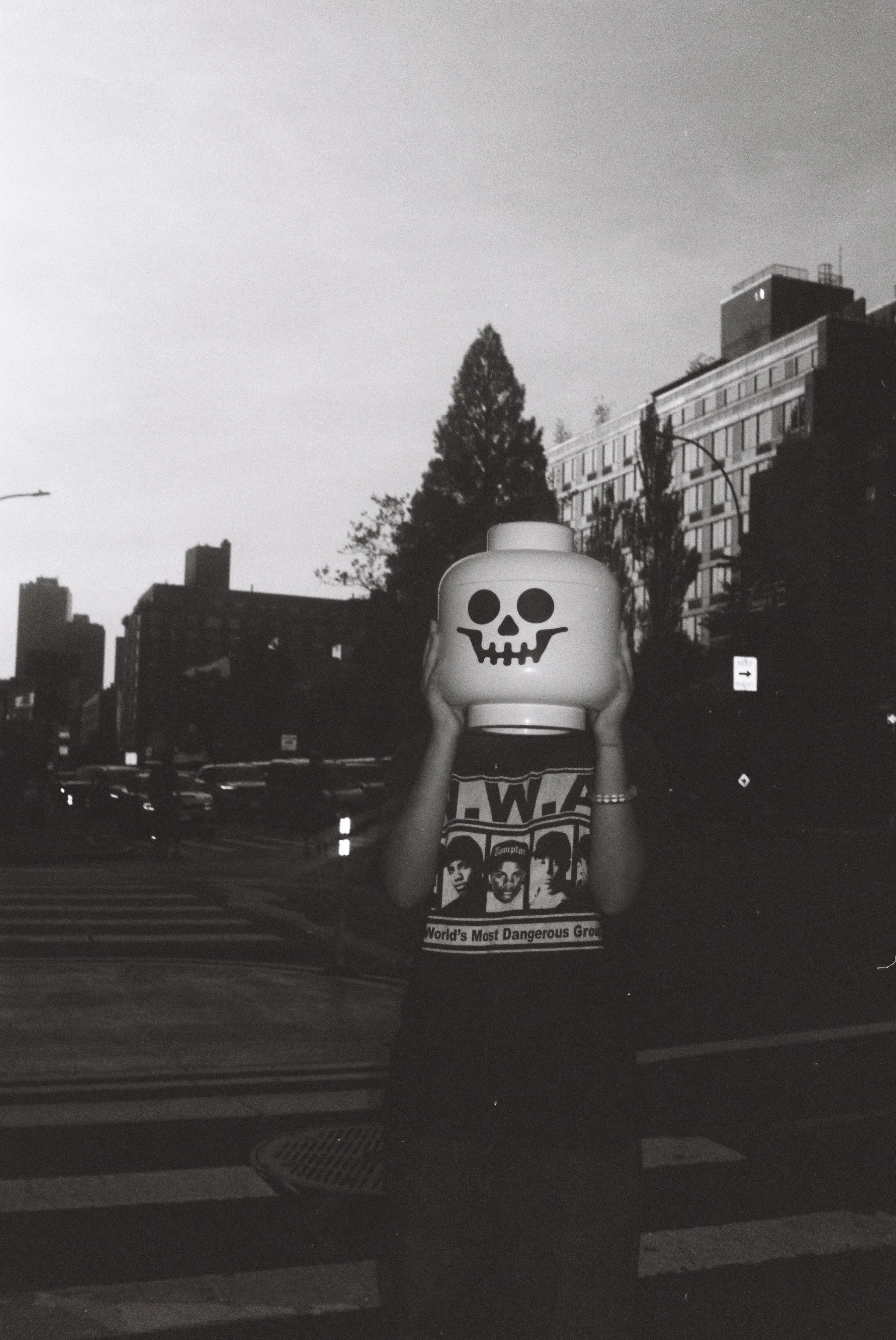 Person standing outdoors holding a lantern with a skull face design covering their head, wearing a band T-shirt with a photo of The Beatles and the text "World's Most Dangerous Group," in an urban setting during dusk or night.