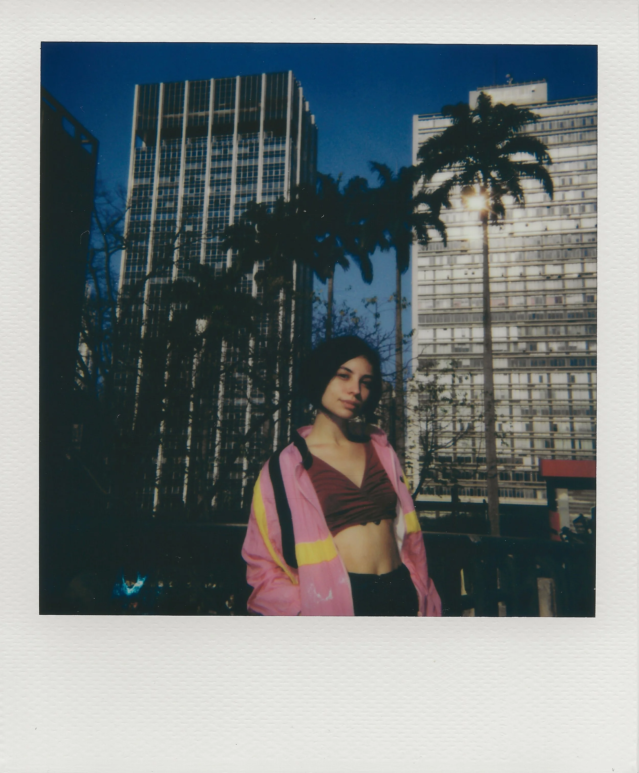 A woman with dark hair wearing a pink jacket and a maroon crop top standing outdoors with tall buildings and palm trees in the background.