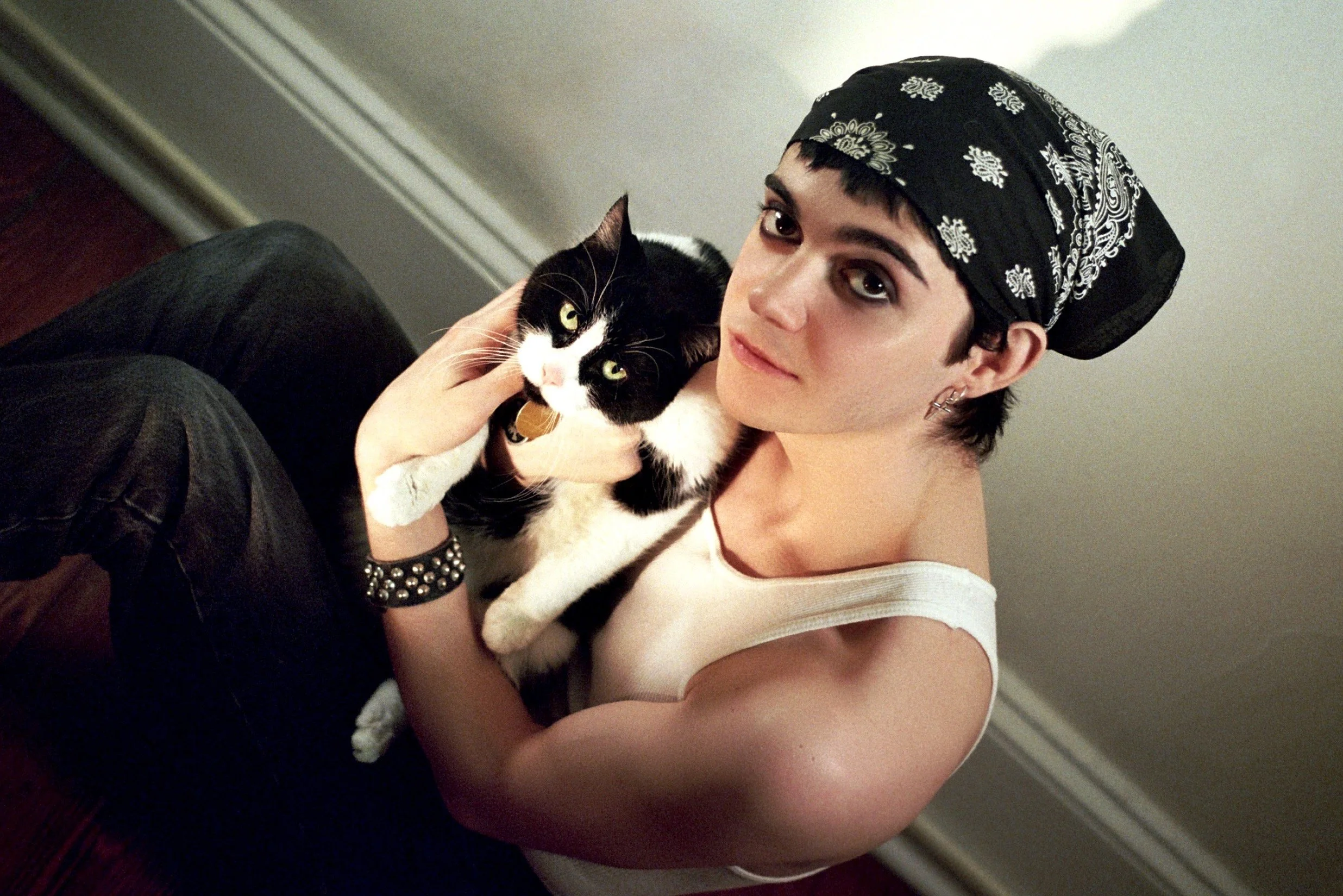 A person with short black hair, wearing a black bandana with white patterns, holding a black and white cat while sitting on the floor indoors.
