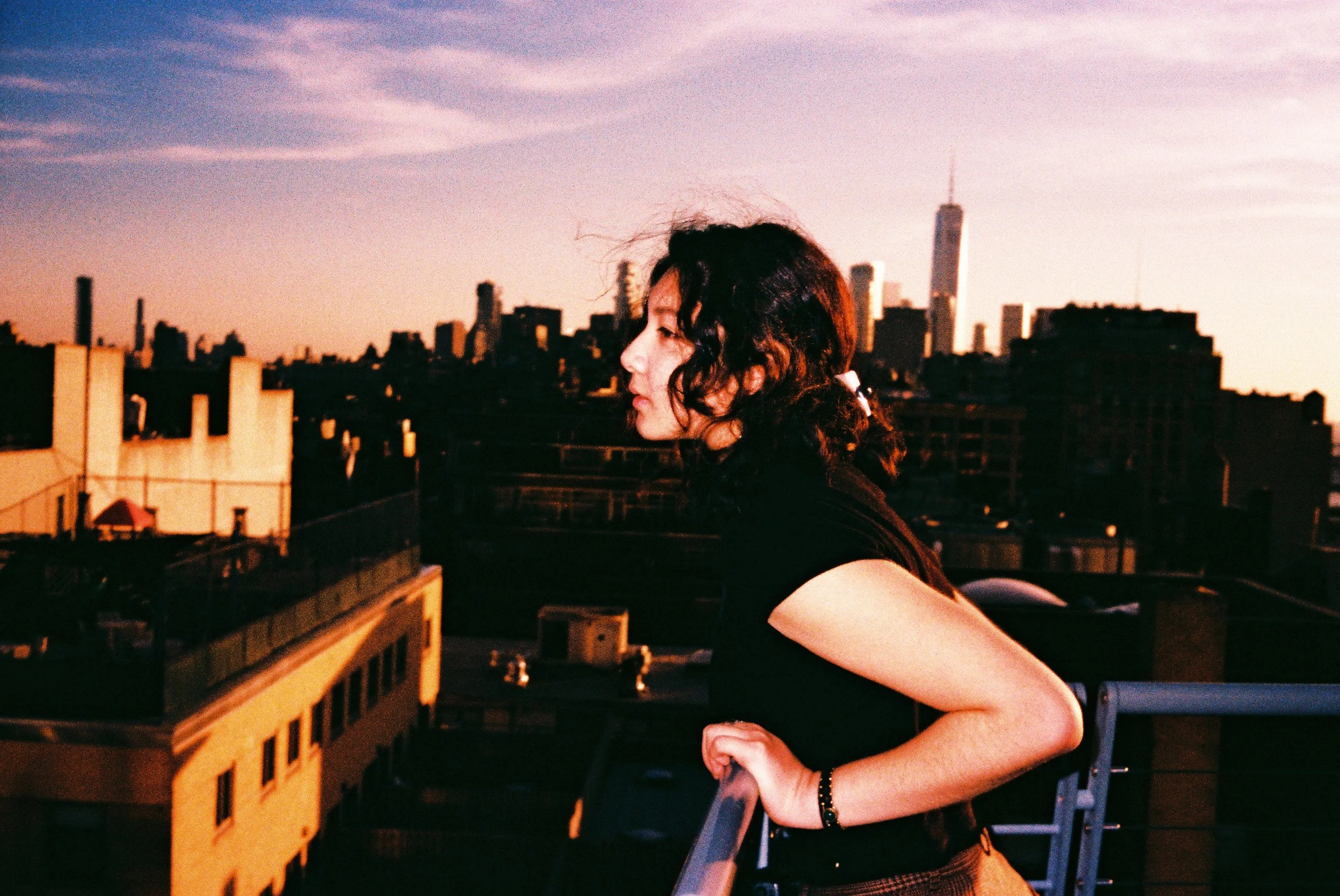 A woman with dark curly hair and a black shirt leans on a rooftop railing, looking to the left, with a city skyline and sunset sky in the background.