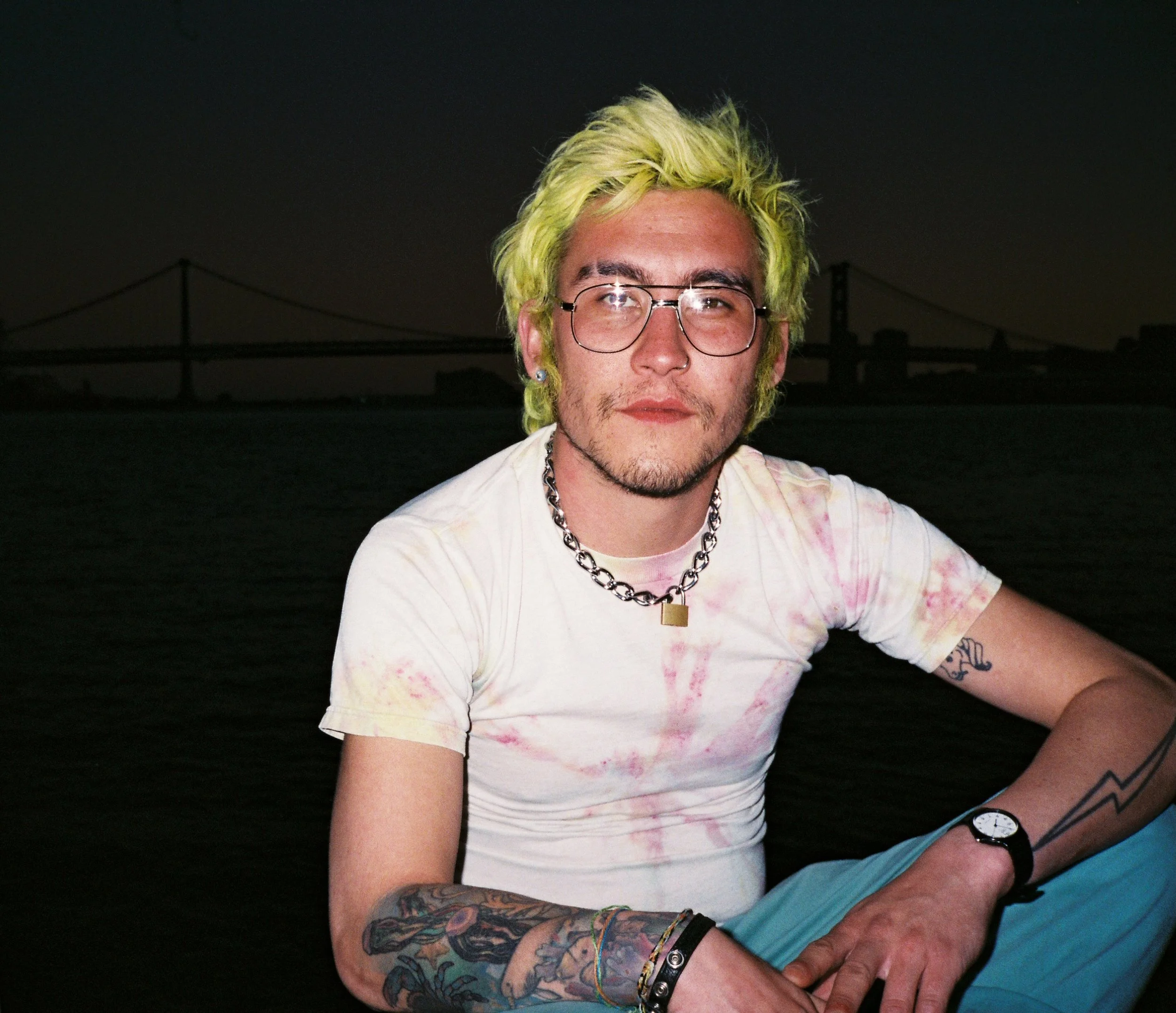 A young man with bright yellow-green hair, wearing glasses, a white tie-dye t-shirt, a silver chain necklace, and watch, sitting outdoors at dusk with a bridge silhouette in the background.