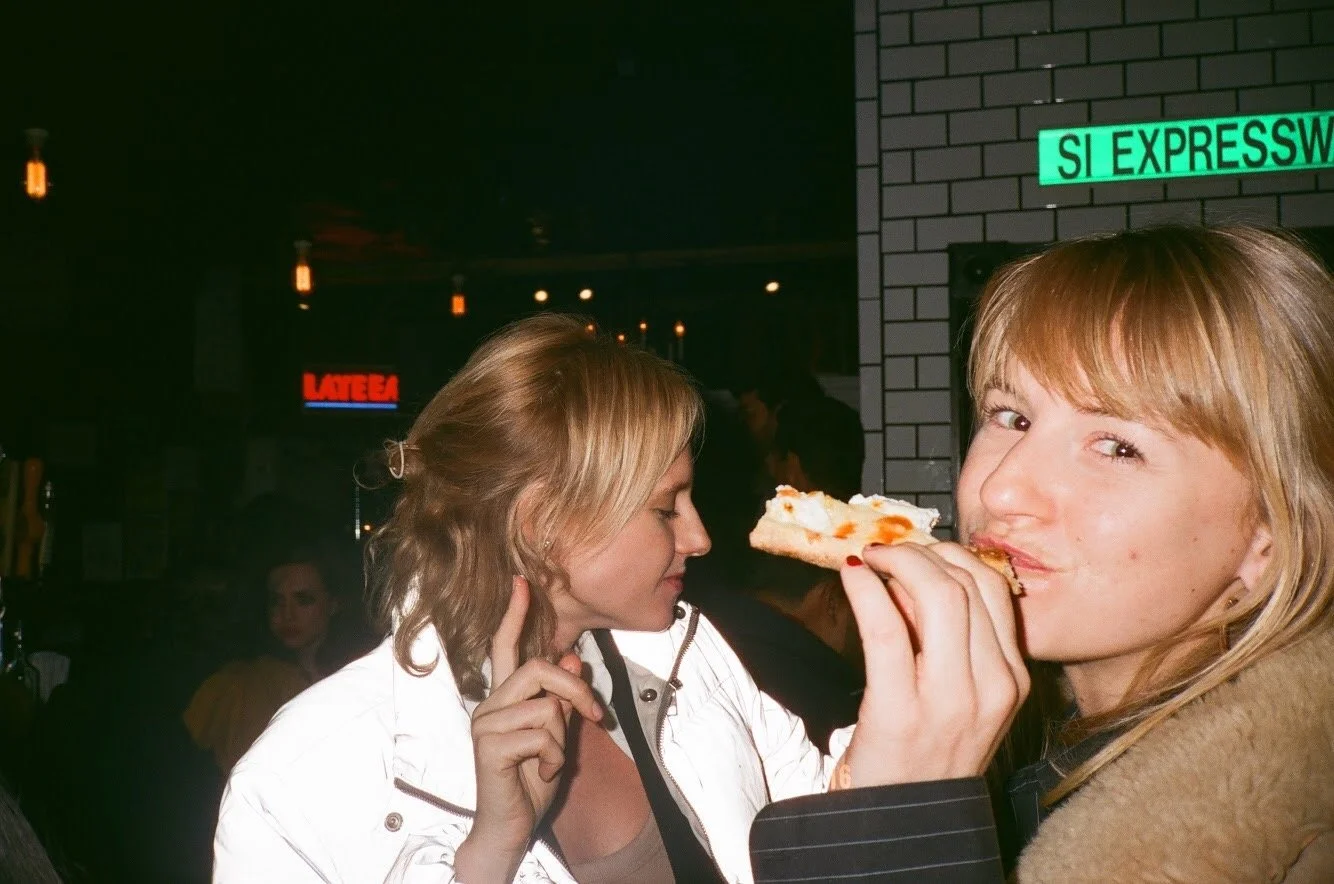 Two young women in a bar or restaurant, one of them eating a slice of pizza, with a dimly lit background featuring hanging lights and signs.