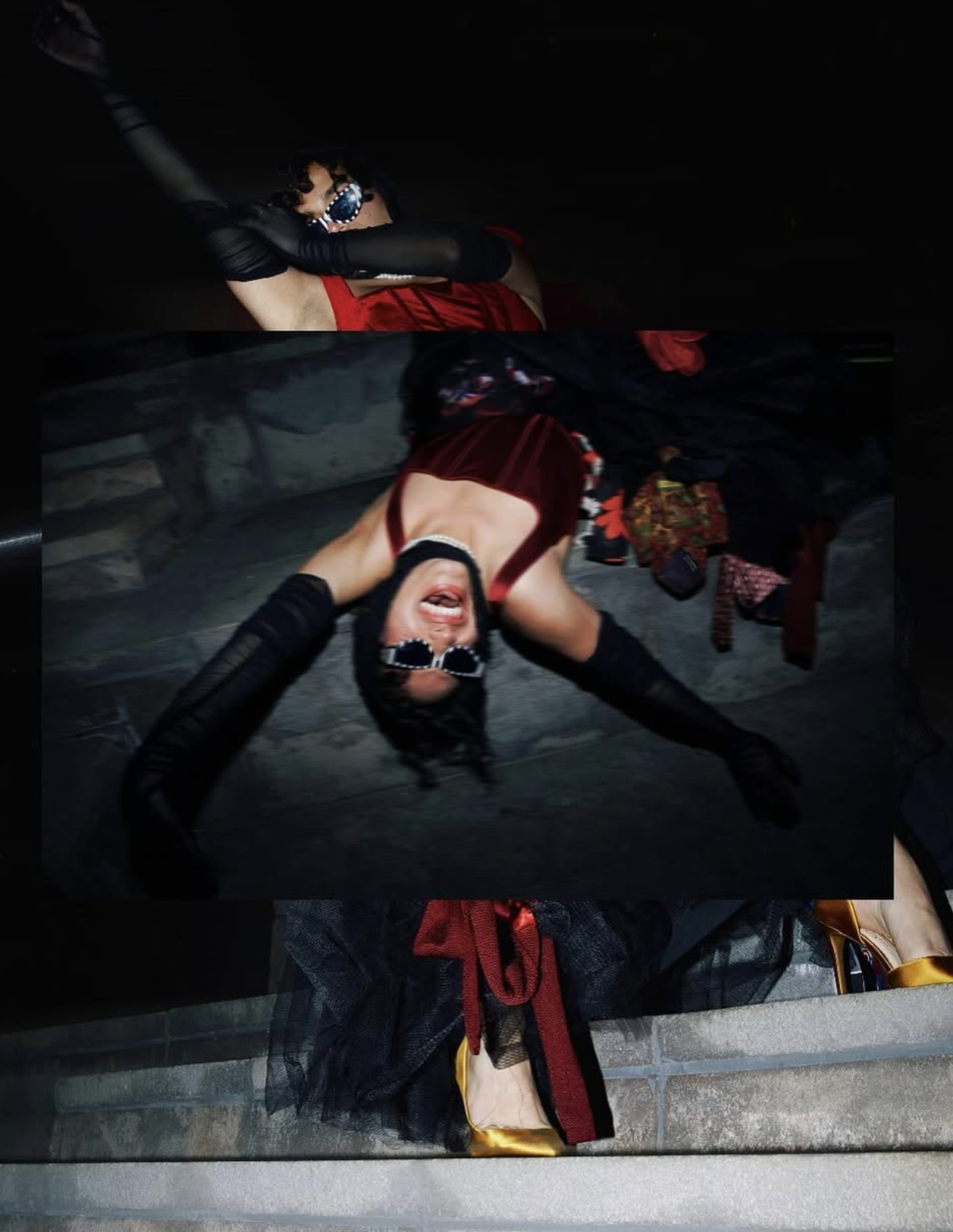 A woman dressed in a red and black outfit with long black gloves, wearing sunglasses, lying on the ground, smiling widely, with a chaotic background, other people's legs and shoes visible. Editorial lifestyle with styling by Louis.