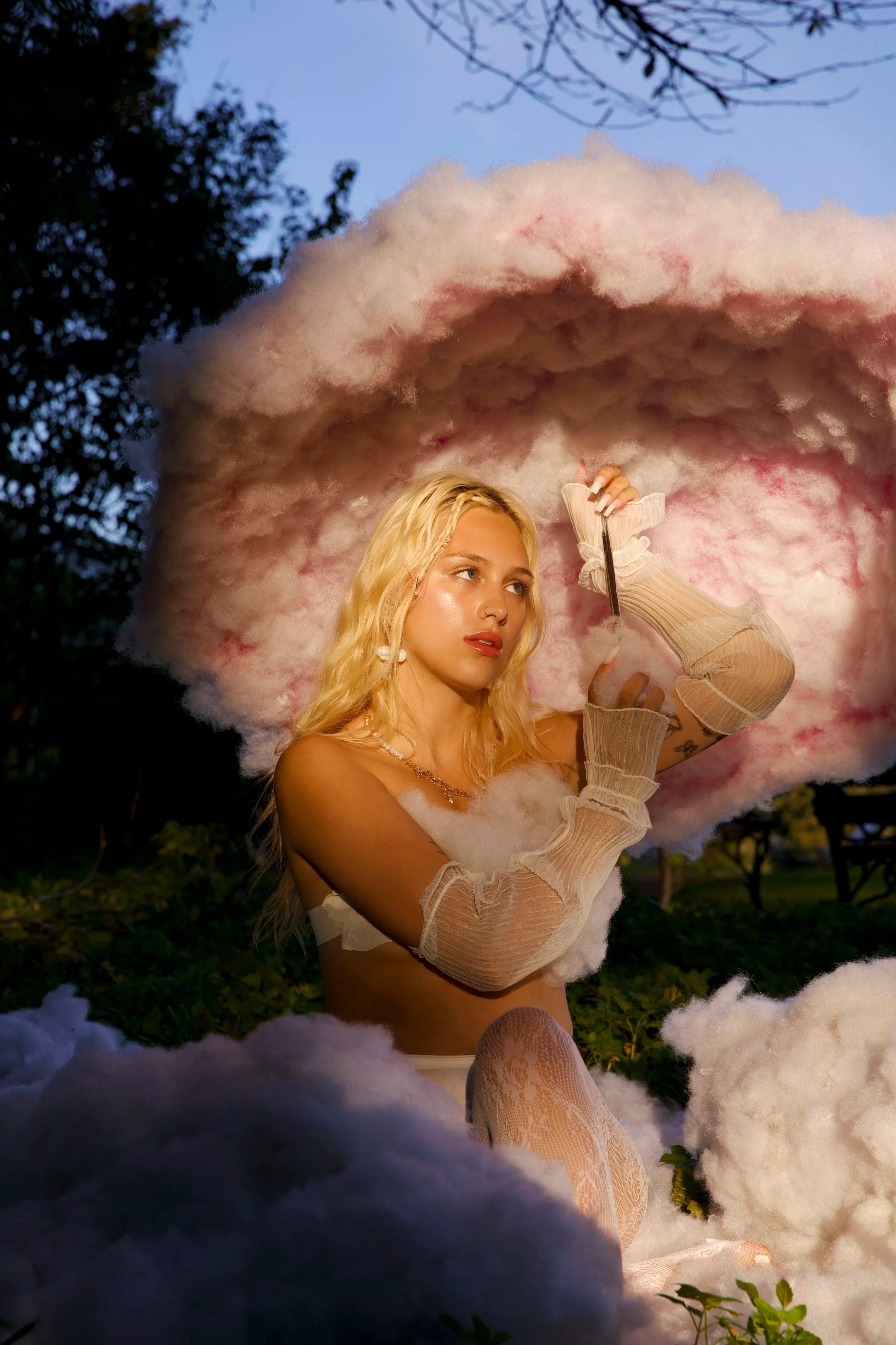 A woman with blonde hair and light skin is surrounded by pink and white cotton candy clouds outdoors during dusk, holding a cotton candy stick, wearing sheer peach-colored gloves, a white top, and lace patterned stockings.