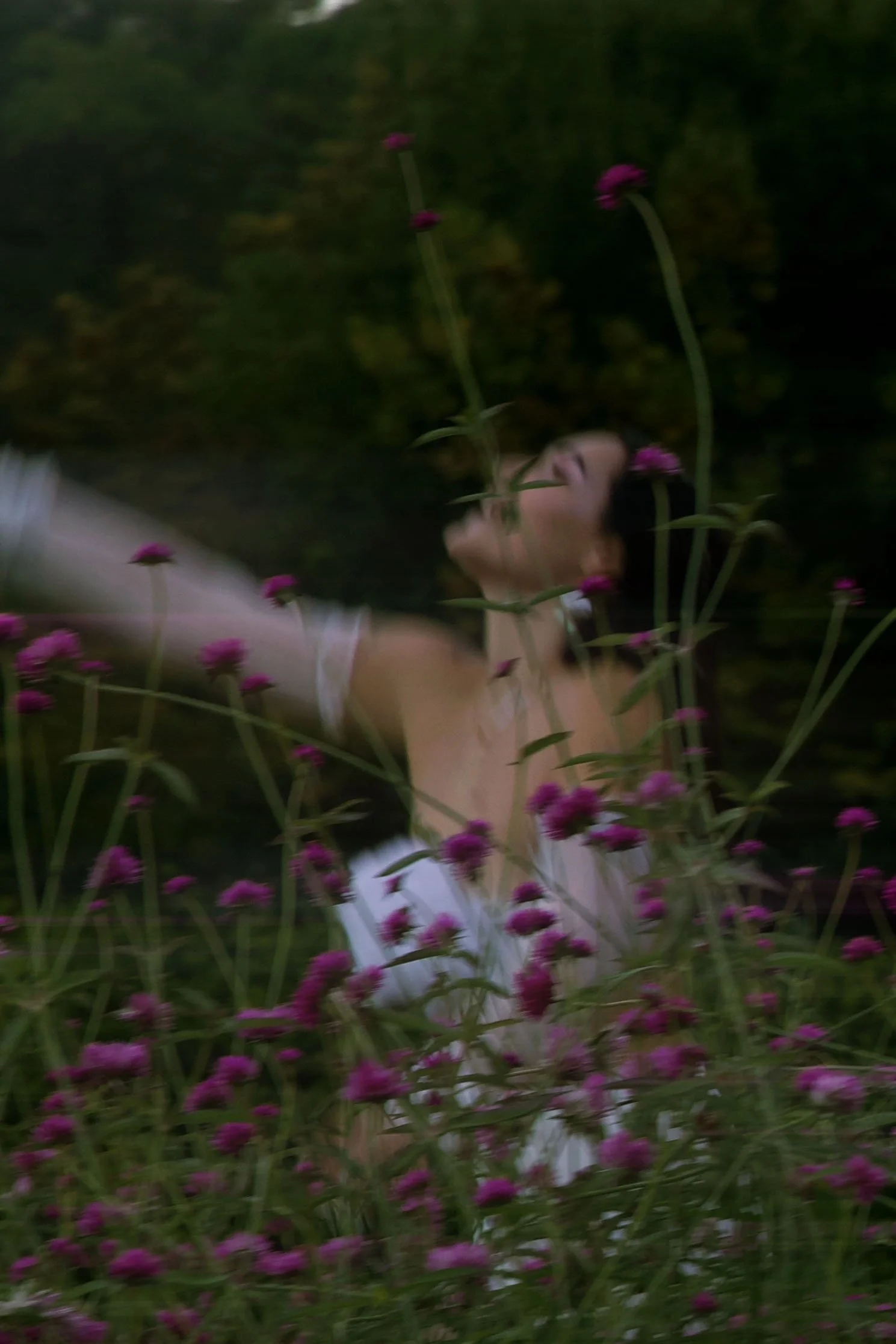 A woman with dark hair wearing a white dress, surrounded by pink flowers, appears to be dancing with her arms outstretched in a garden or natural setting. Witchy lifestyle photoshoot.