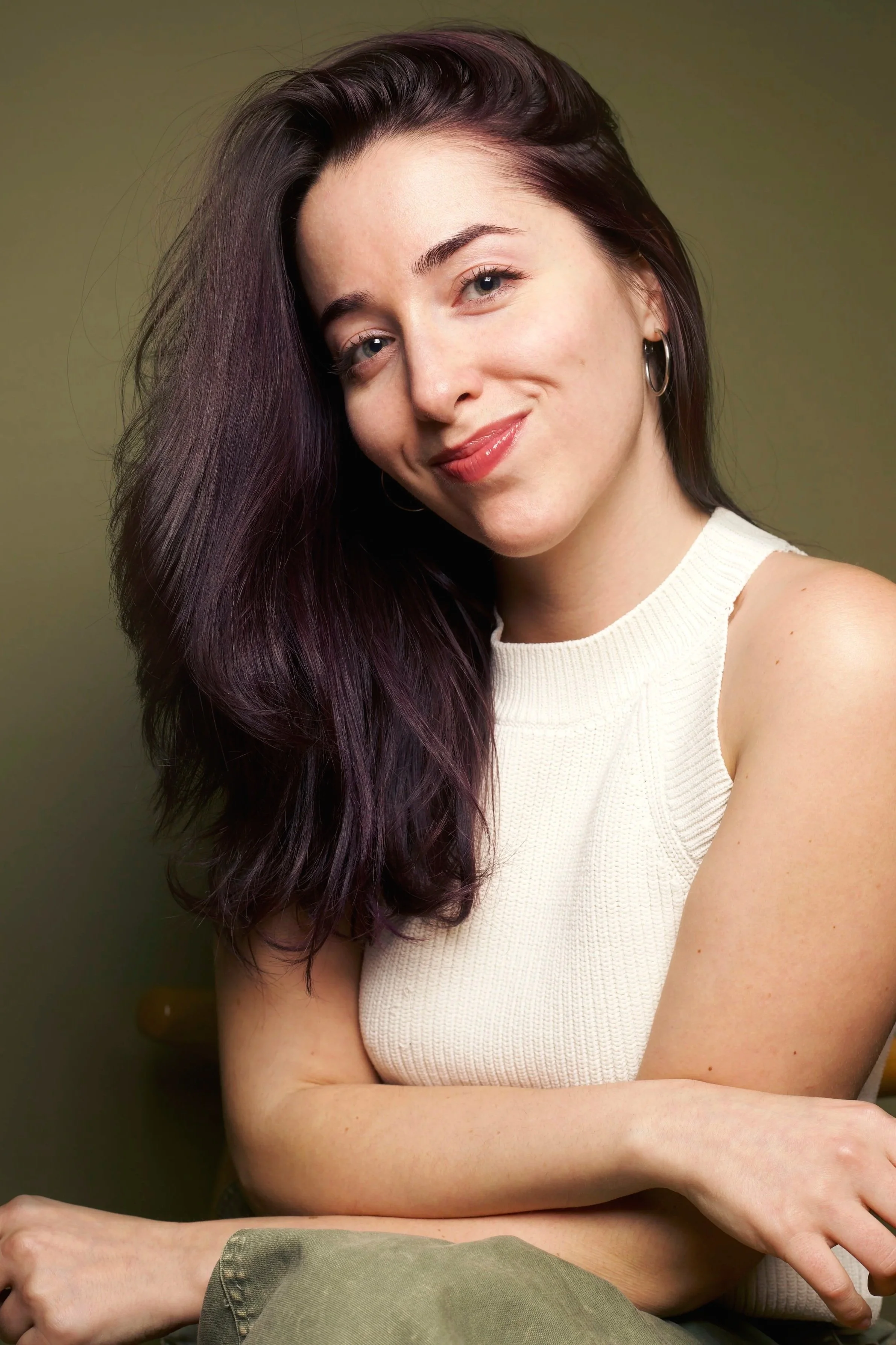 A young woman with long, dark purple hair, smiling, wearing a sleeveless white knit top, earrings, and sitting with crossed arms against a muted green background. Self portrait of miss cola. Alex Colavecchio.