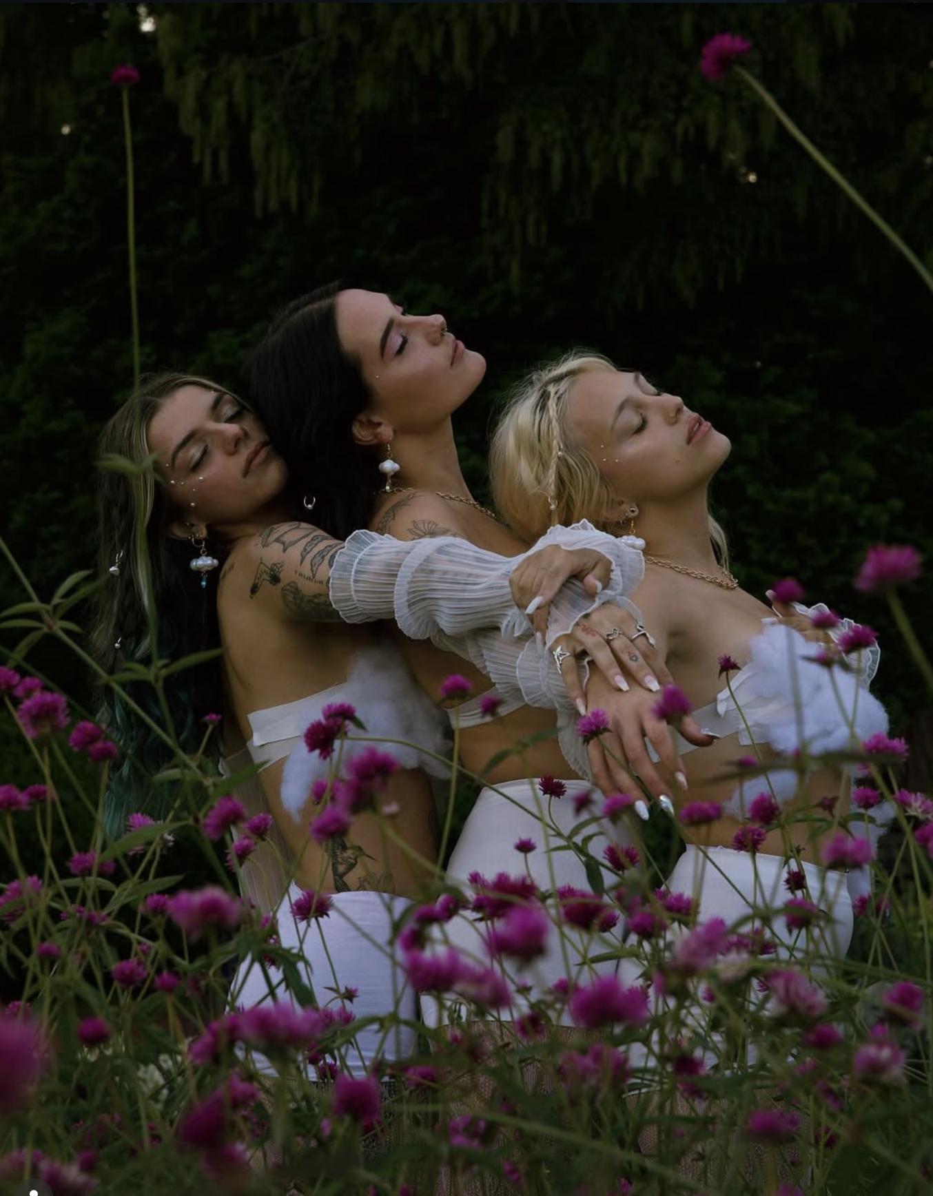 Three women with tattoos and jewelry, dressed in white, are embracing in a field of pink flowers with a dark green forest background, with their eyes closed and faces relaxed. Witchy garden lifestyle shoot.