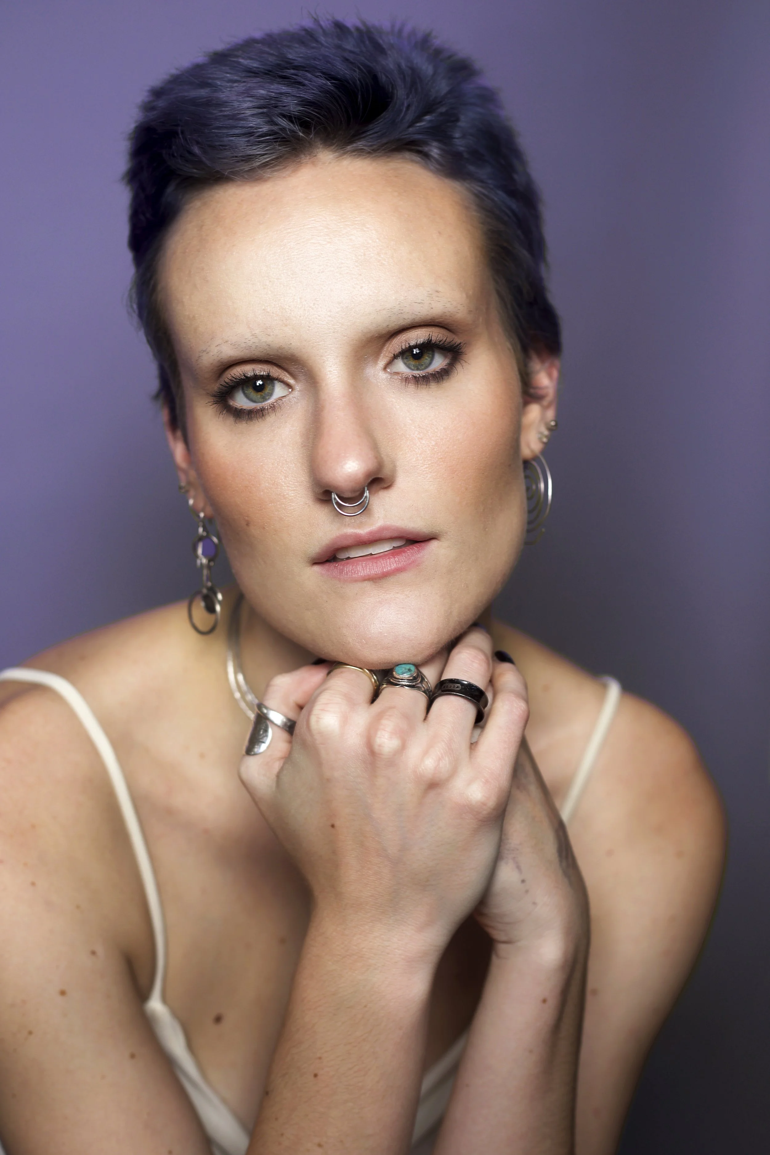 Close-up portrait of a woman with short dark hair, multiple rings, earrings, and septum piercing, with hands resting on her chin, against a purple background.