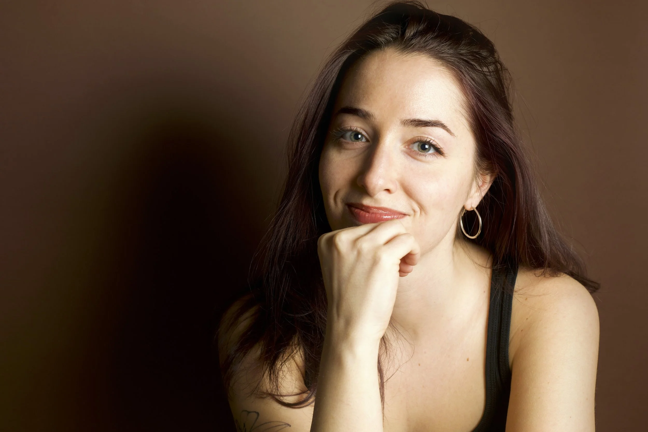 Portrait of a smiling young woman with long dark hair, wearing hoop earrings, resting her chin on her hand against a brown background. Self Portrait of miss cola. Alex Colavecchio.