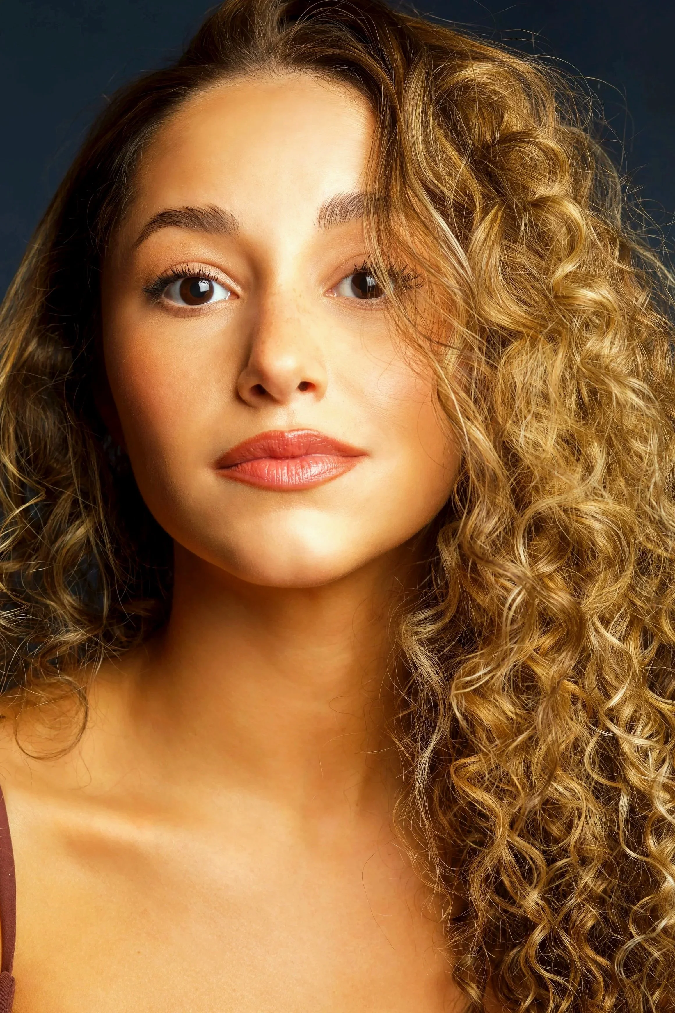 Close-up portrait of a young woman with curly blonde hair, light makeup, and a slight smile, against a dark background.
Striking headshots.