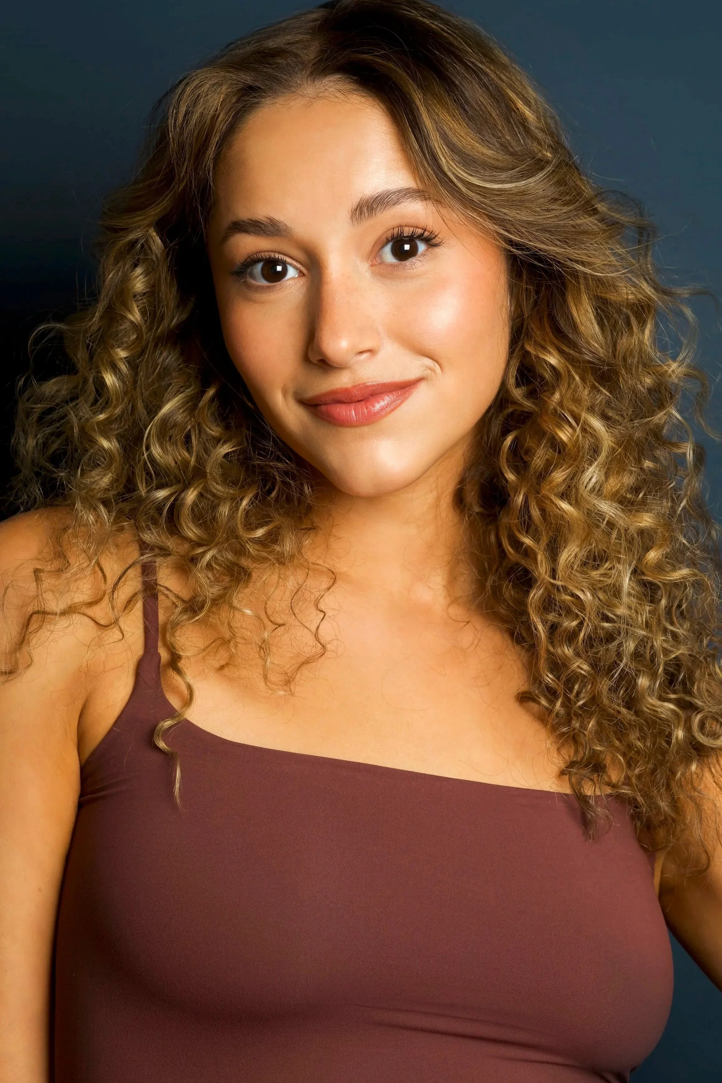 A young woman with curly hair and a warm smile poses against a dark background, wearing a sleeveless maroon top.