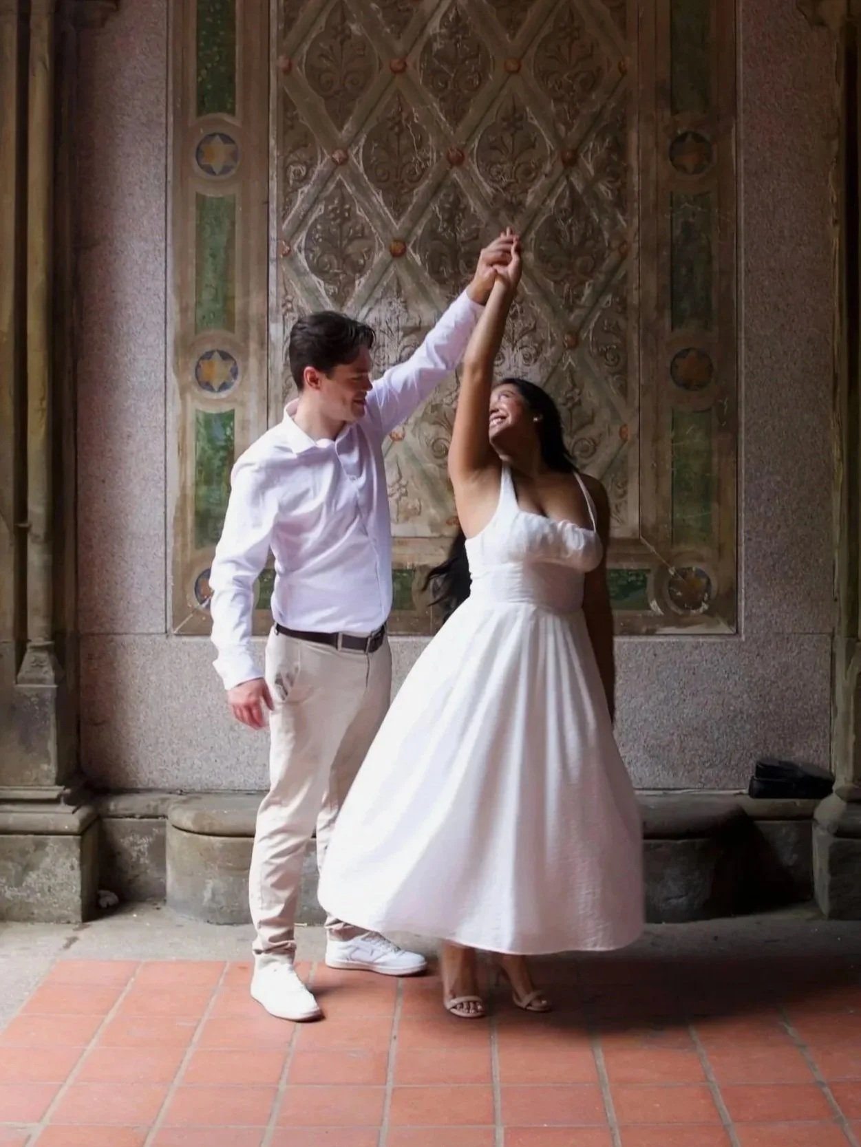 A man and woman in wedding attire dancing together indoors, with the man holding the woman's hand high and both smiling.
